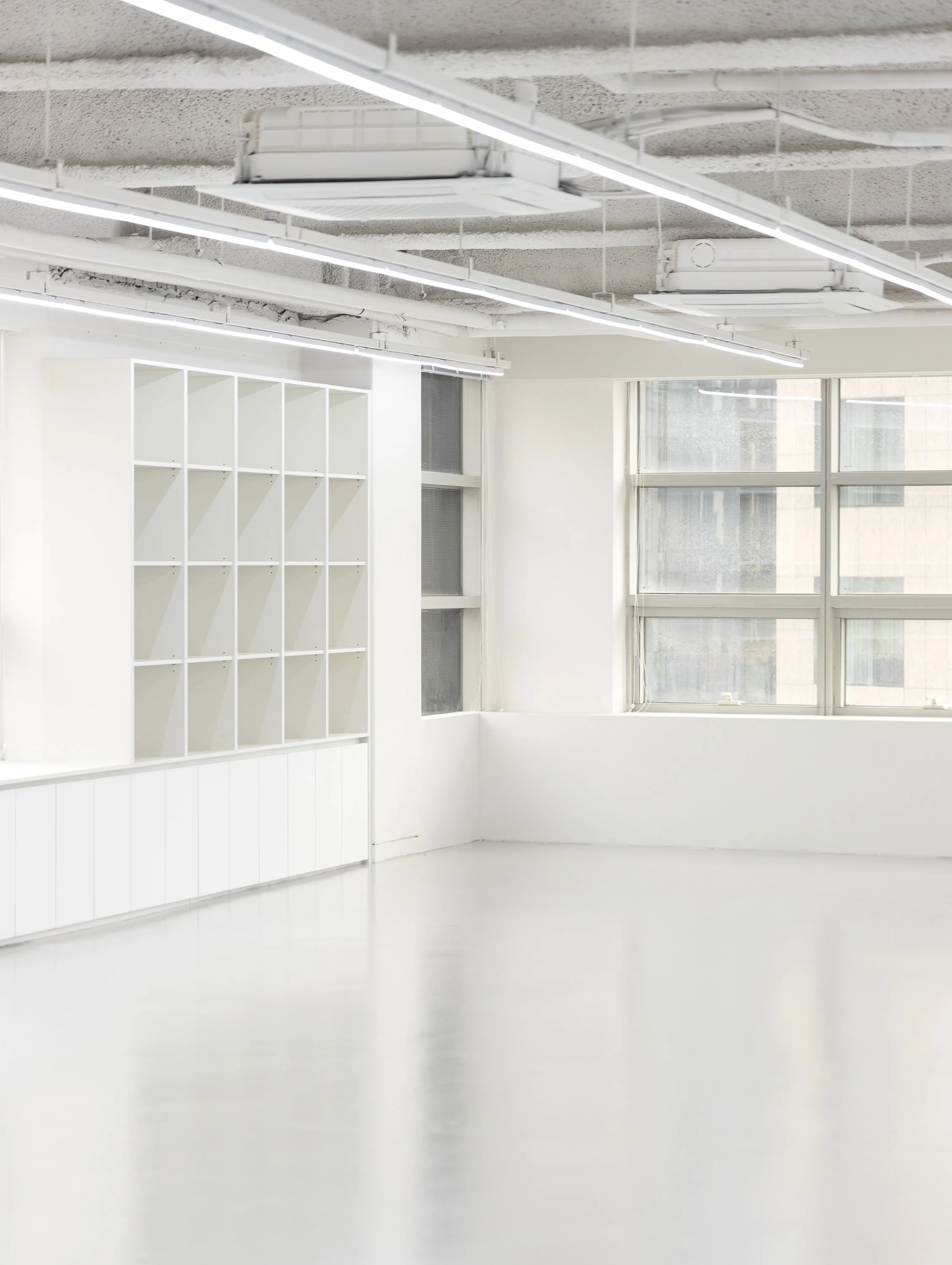 Empty white room with large windows, white shelving unit, and exposed ceiling with lighting fixtures and ductwork.