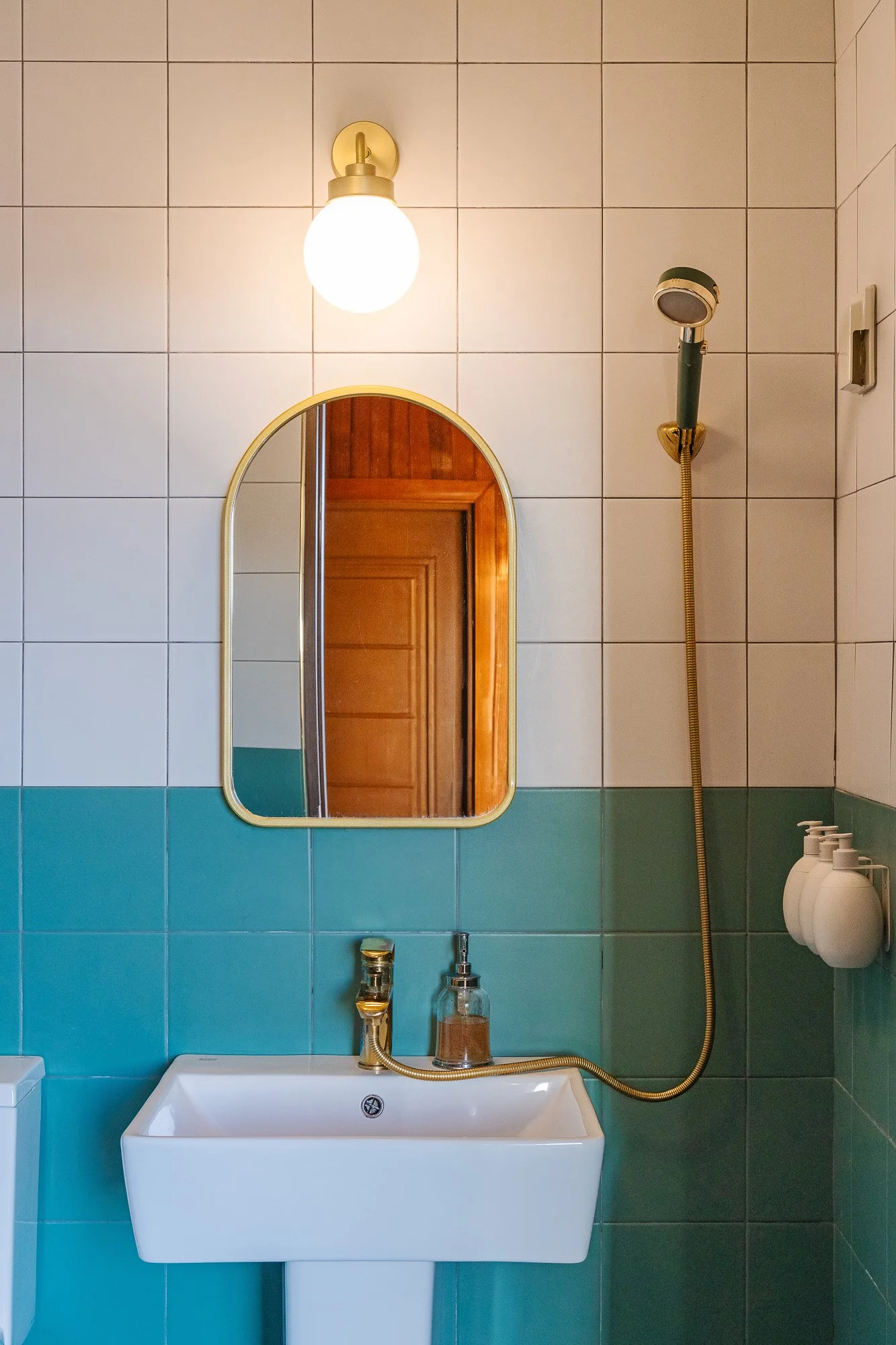 Bathroom with beige and teal tiled wall, white sink, gold faucet, soap dispenser, mirror, wall-mounted light, and shower head on a flexible hose.