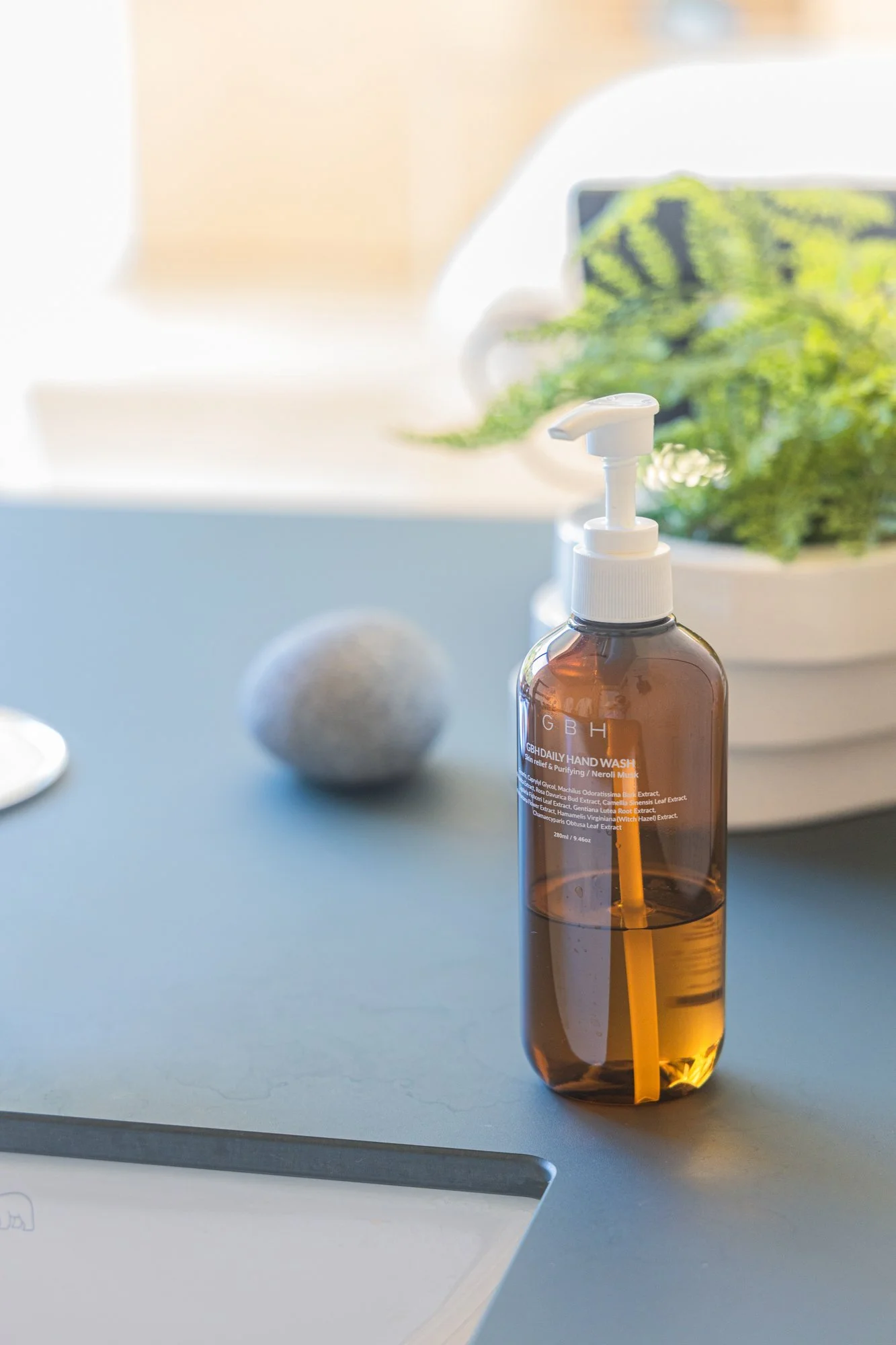 A hand soap dispenser on a desk with a grey ball, a white notepad, and a planter with green plants in the background.