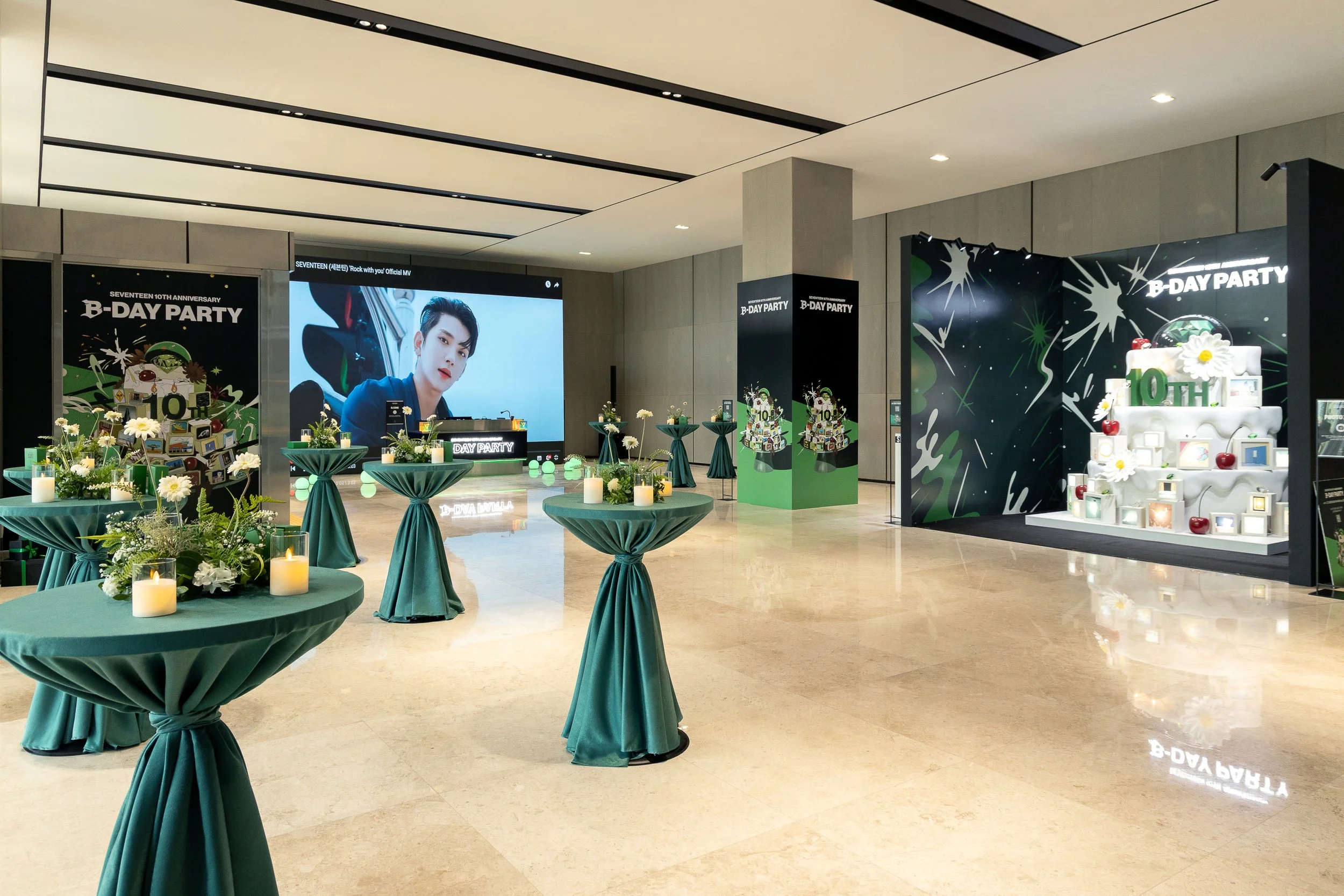 A decorated event space celebrating SEVENTEEN's 10th anniversary with a large screen showing a member of the group, floral arrangements, candles, and a tiered cake with the number 10 and flowers.