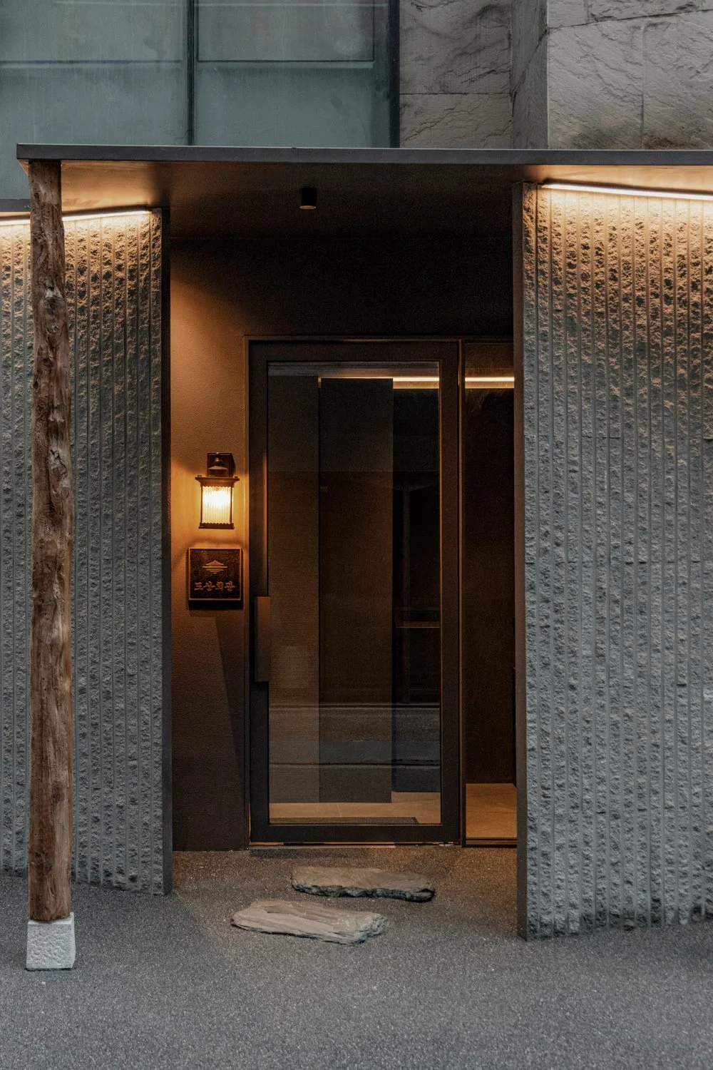 Modern building entrance with a glass door, illuminated wall-mounted lantern, and textured concrete walls, with a wooden support beam on the side.