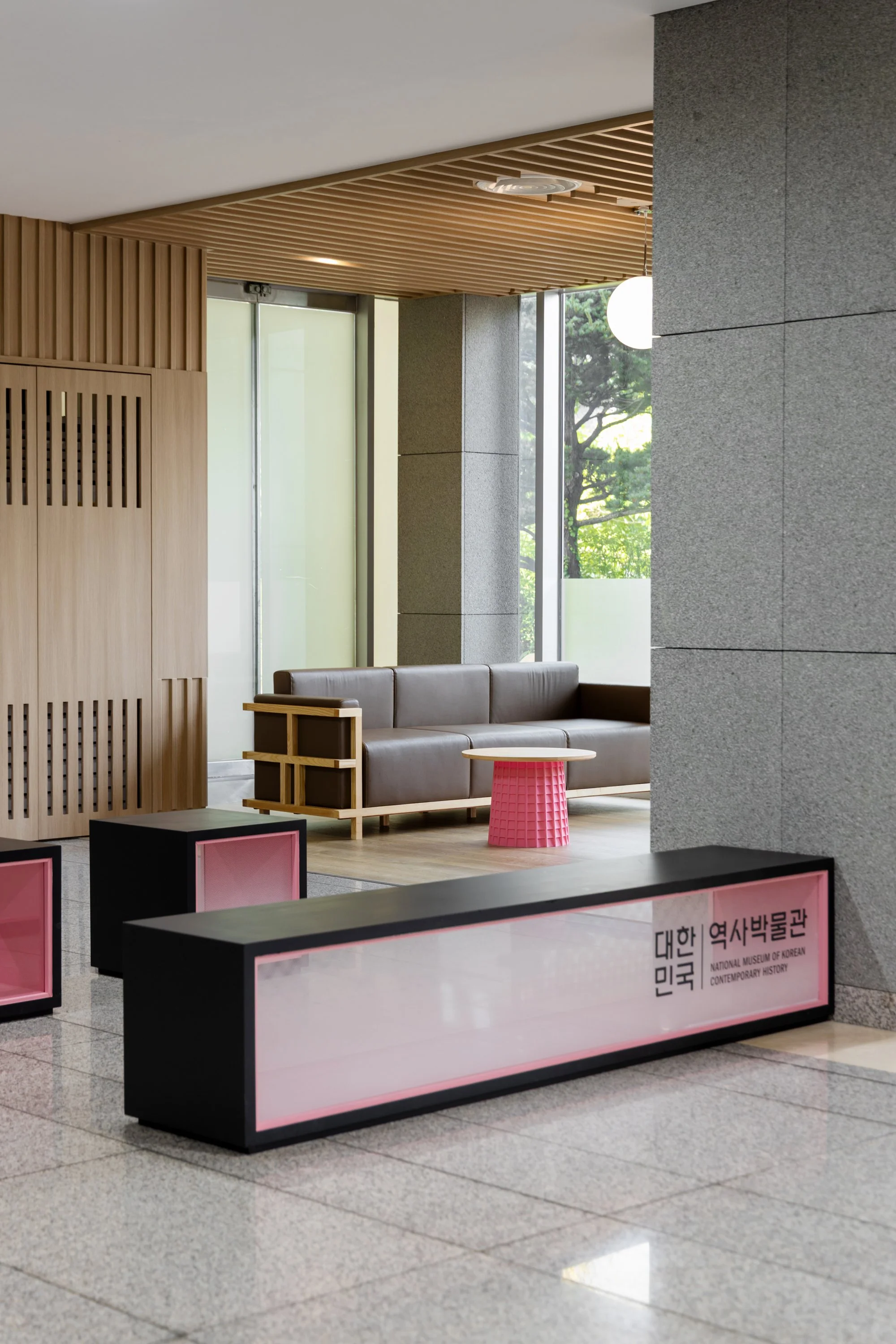 Interior of a modern museum lobby with a brown leather couch, a pink side table, and a digital sign in Korean and English for the National Museum of Korean Contemporary History.