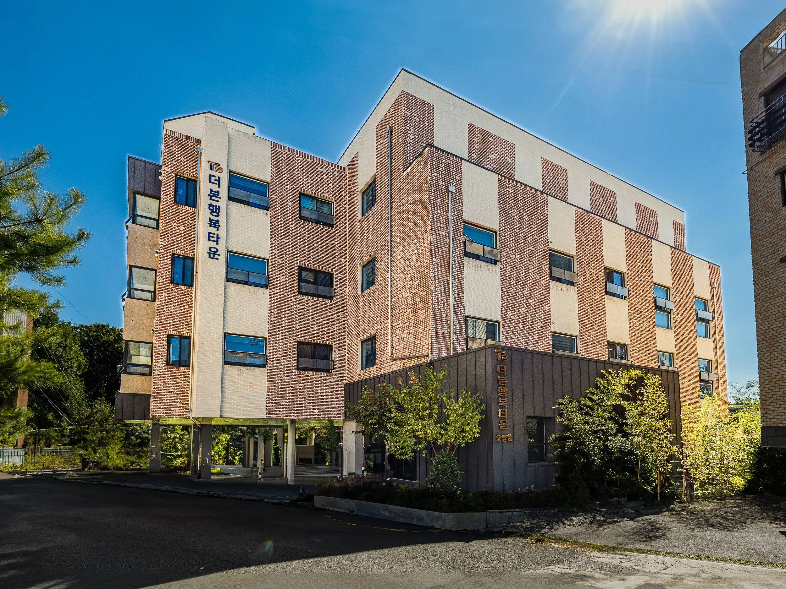 A multi-story residential building made of brick and concrete, with several windows and trees in the front yard, under a bright blue sky with sunlight.