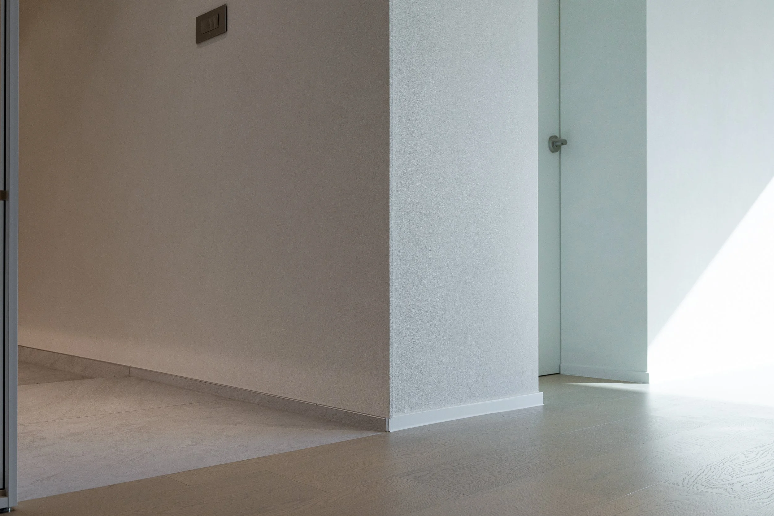 An interior view of a corner with a hallway on the left and a door on the right, featuring minimalist decor, light-colored walls, and flooring.