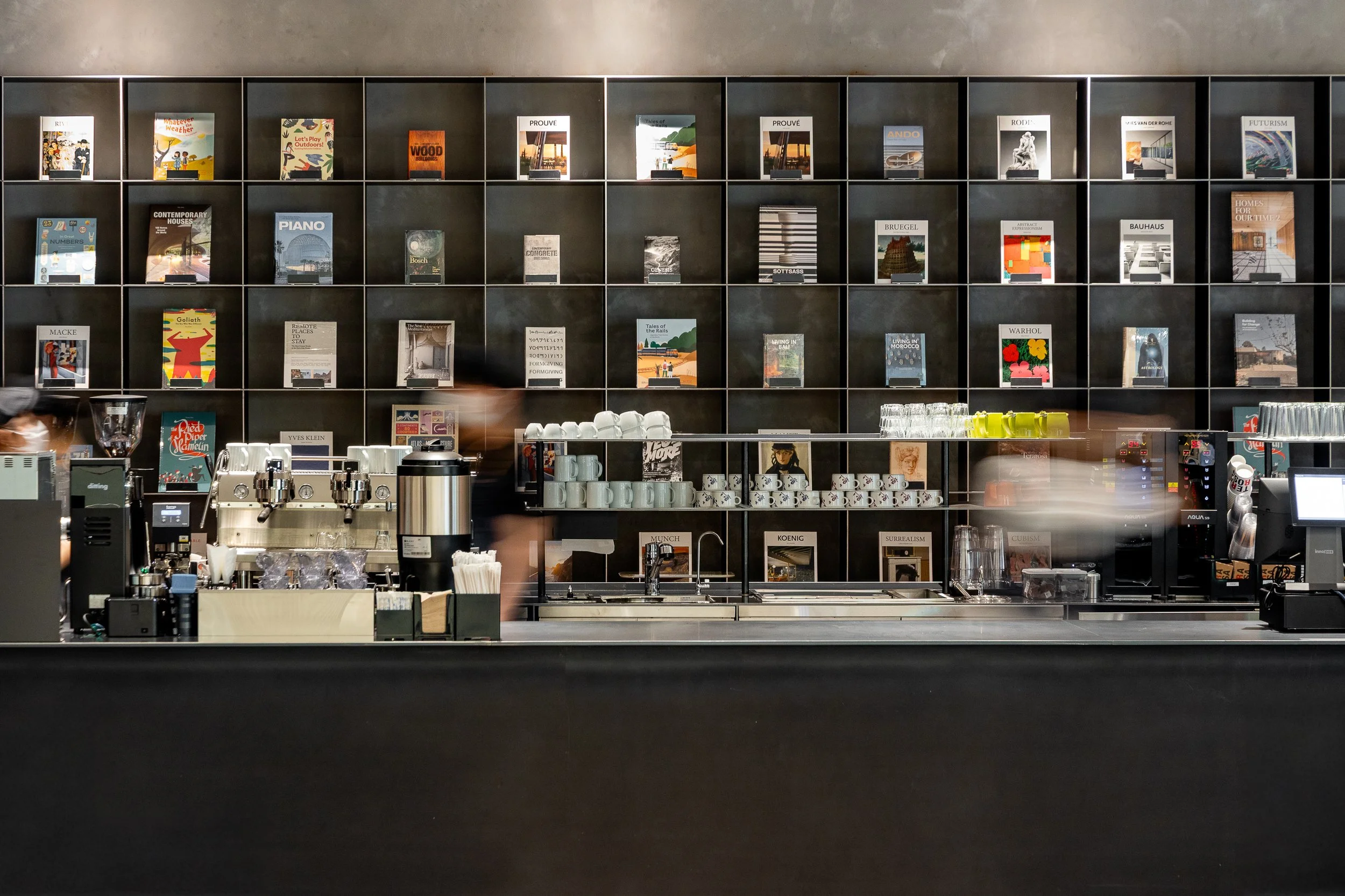A modern coffee shop with a black wall filled with books and magazines on shelves behind the counter. The counter has a coffee machine, cups, and glassware, with blurry staff working behind it.
