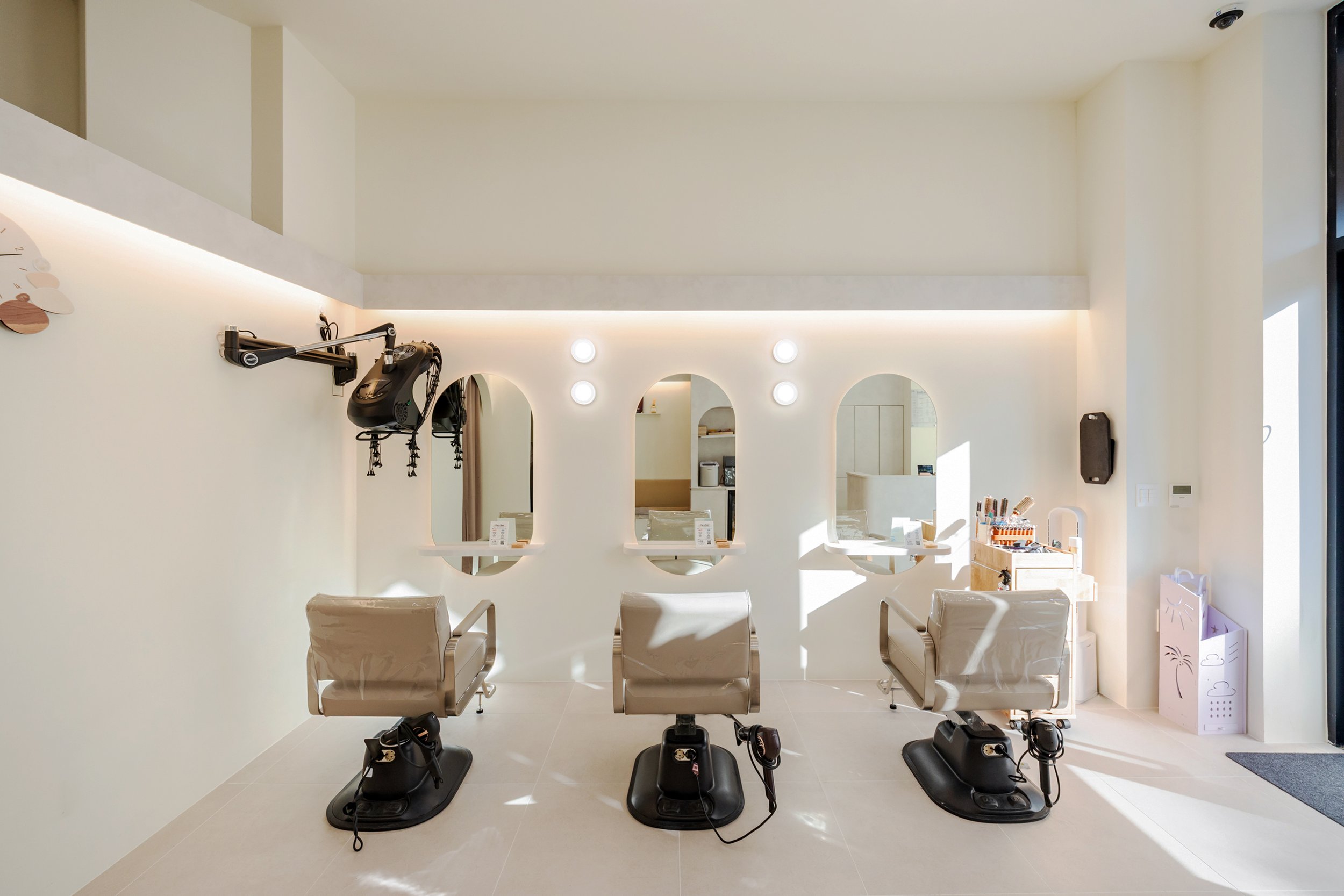 Modern hair salon with three beige styling chairs facing mirrors with arches, sunlight streaming in, and hair styling tools on a small table to the right.