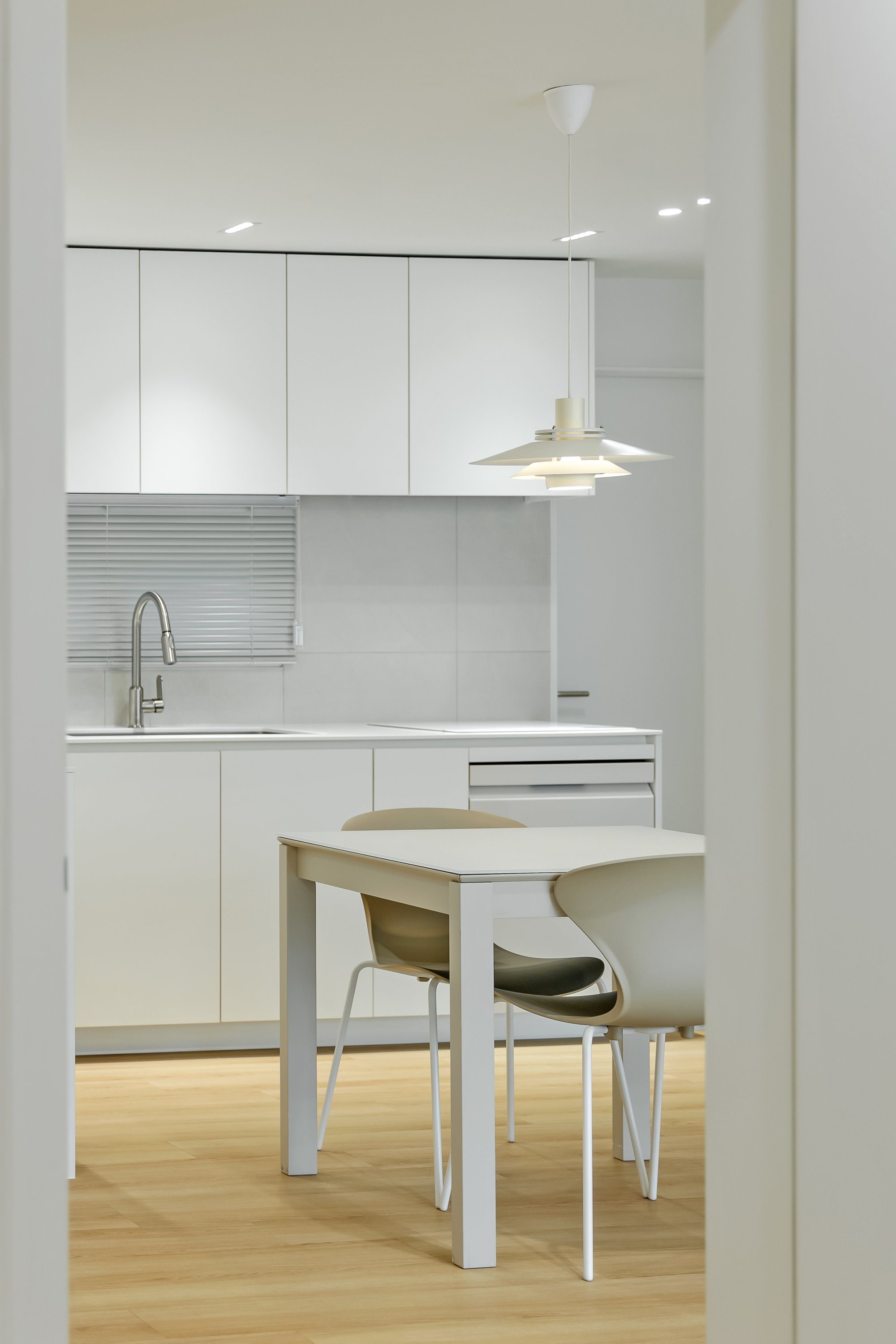 Minimalist kitchen with white cabinets, a sink, a white dining table, and beige chairs, illuminated by a hanging ceiling light, with light wooden flooring.