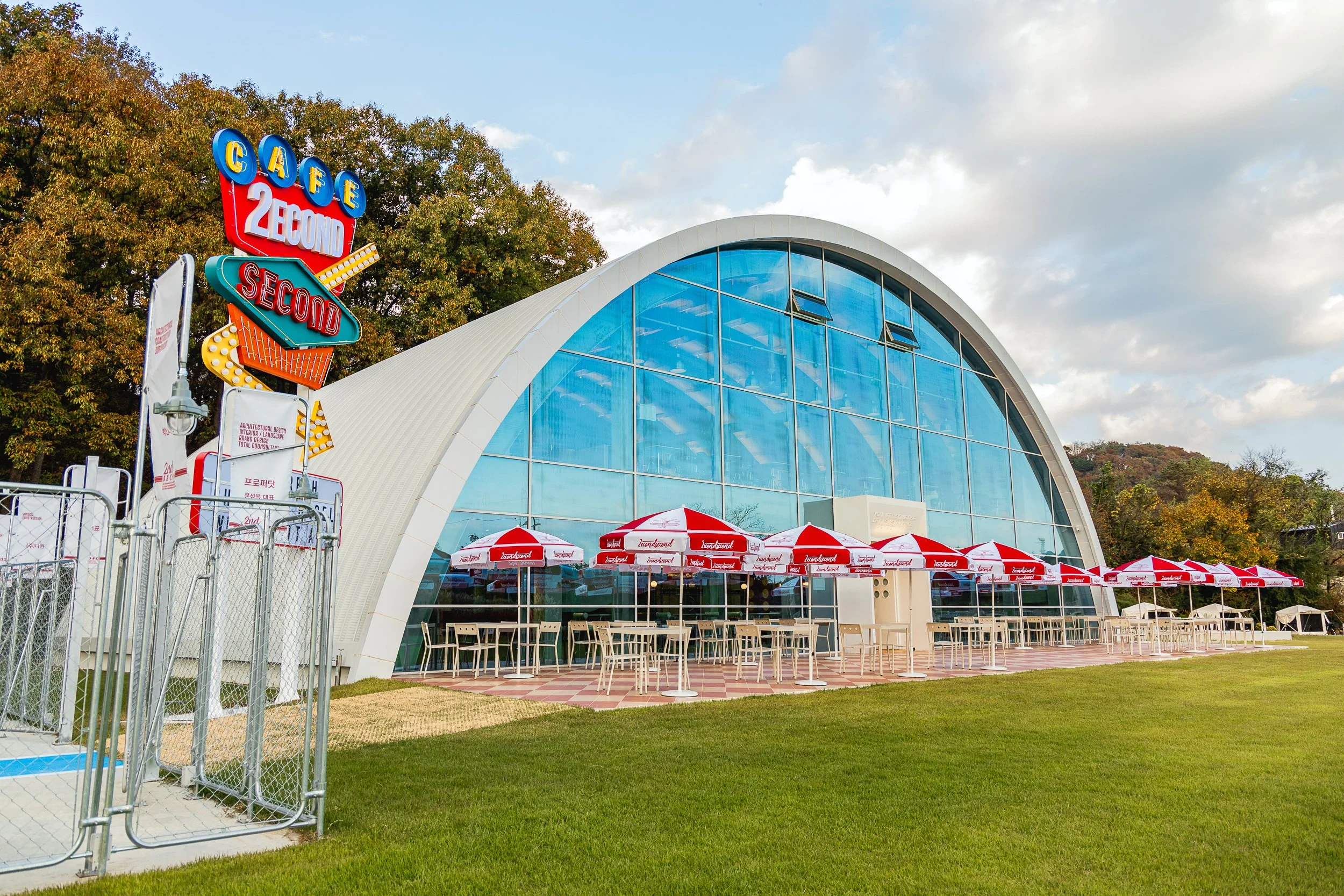 A modern building with a large curved glass front, surrounded by white and red umbrellas and outdoor seating, with a colorful neon sign that reads 'Café 2nd Second', set against a backdrop of trees and partly cloudy sky.