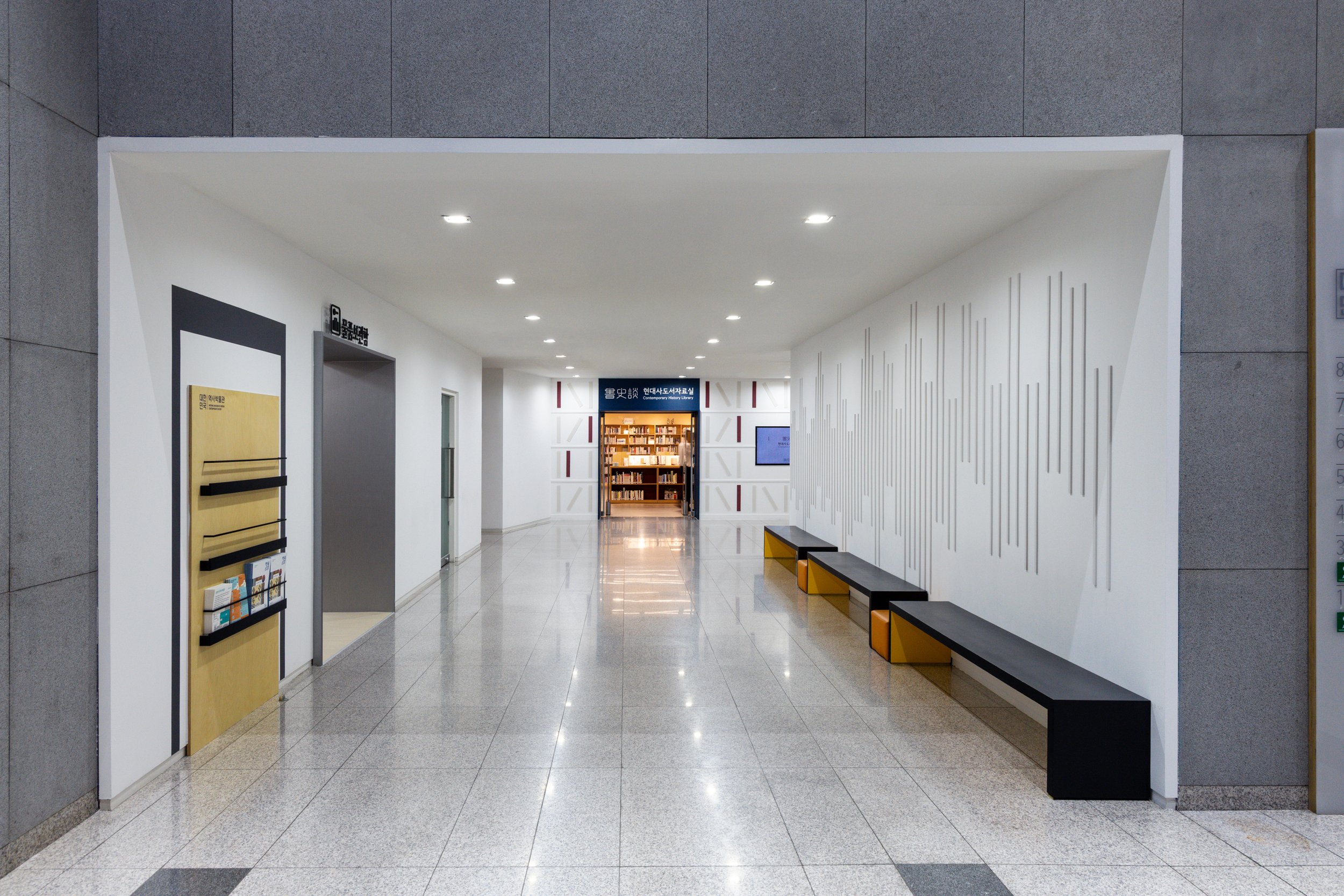 Empty modern library corridor with white walls, gray and yellow benches, and a library at the end.