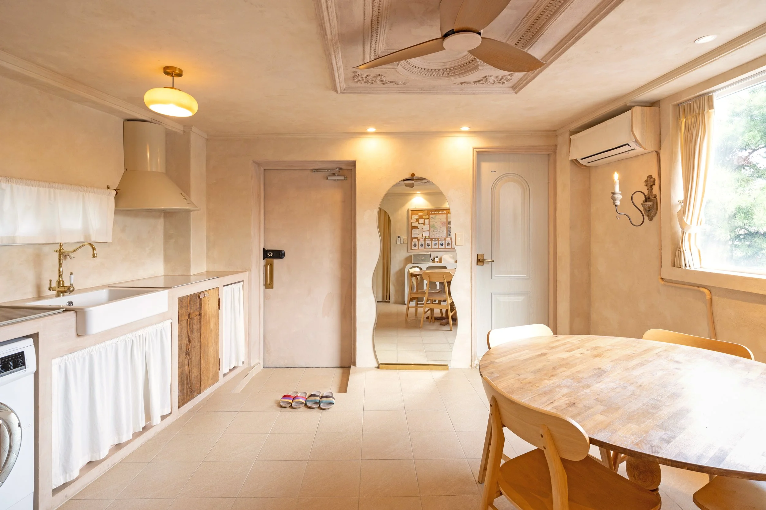 Cozy kitchen and dining area with round wooden table, chairs, a mirror, window with curtains, air conditioning, ceiling fan, slippers on the floor, and warm lighting.