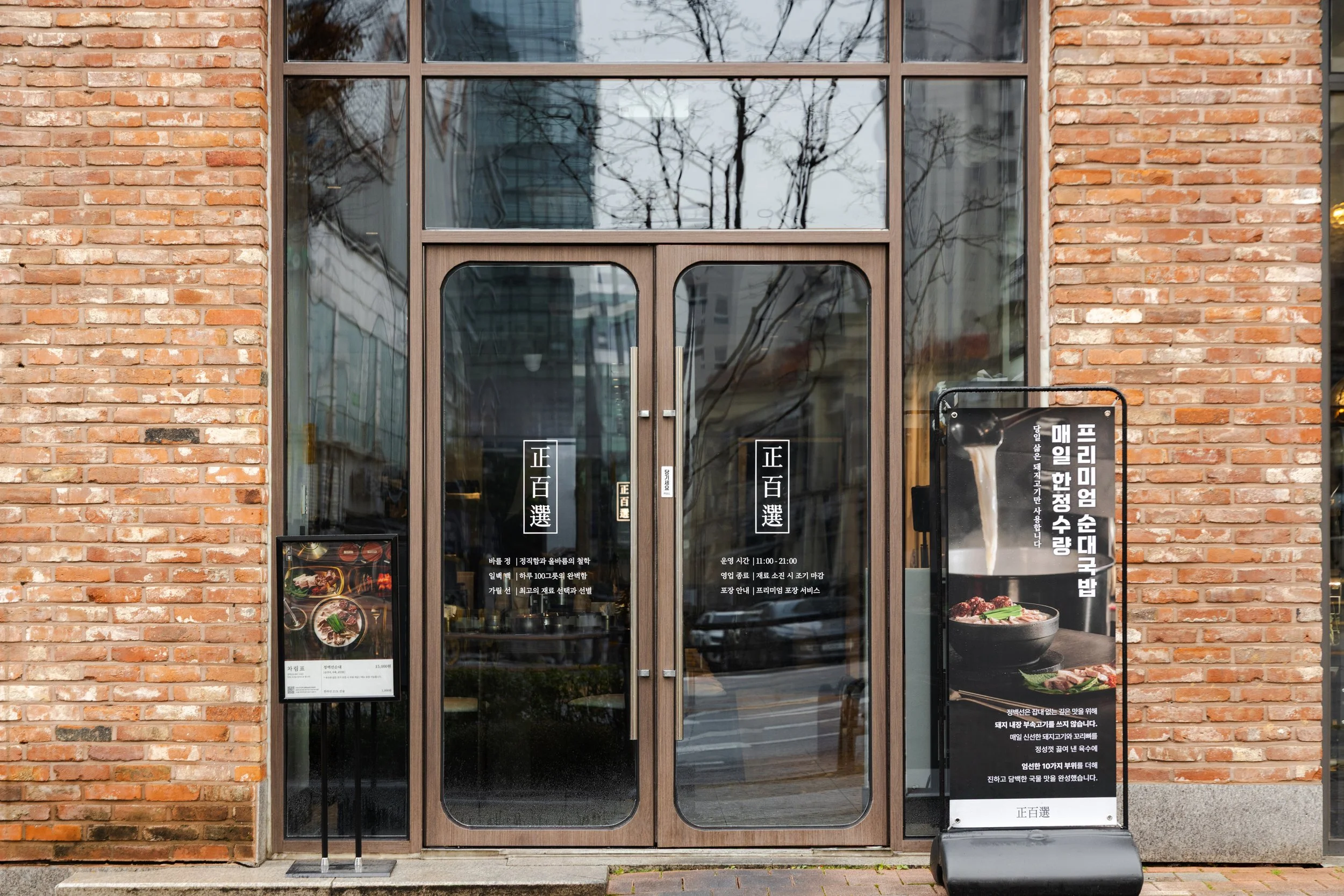The entrance of a Korean restaurant with glass double doors and brick walls. There are signs with Korean text and pictures of food outside the restaurant.
