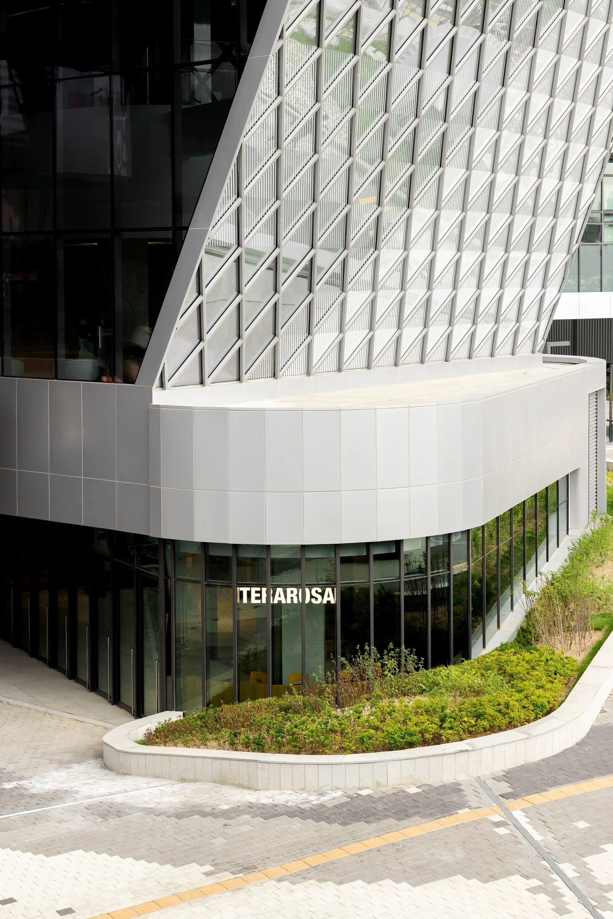 Modern building with large glass windows and a distinctive geometric façade. The ground floor has a sign that reads 'TETAROSA' and is surrounded by landscaped greenery and a paved walkway.