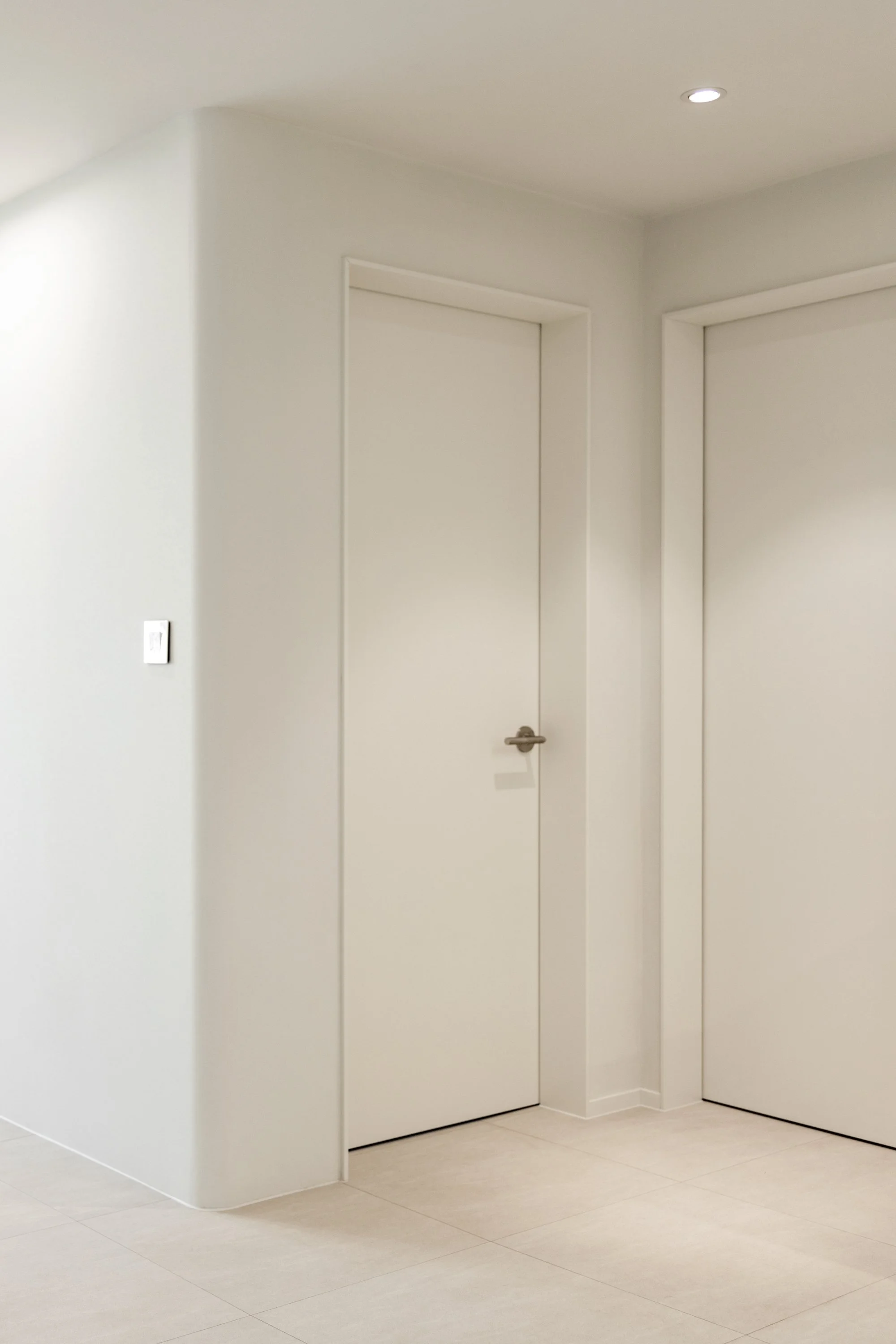 A white interior hallway corner with two closed doors, one on the left and one on the right, and a small section of beige tile flooring.