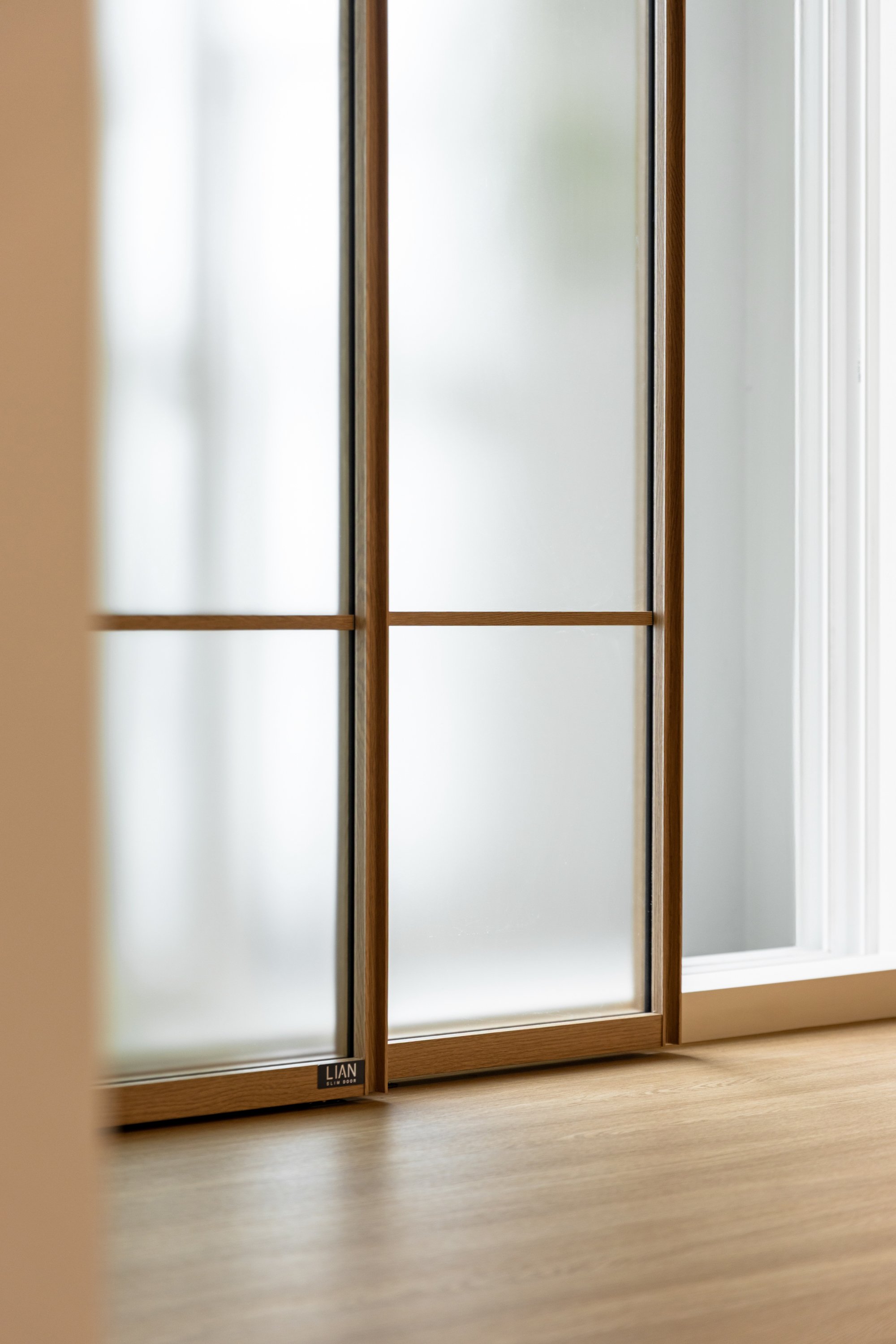 Close-up of a wooden and frosted glass sliding door with a small black label that reads 'LIAN' at the bottom left corner, with white walls and a wooden floor beyond.