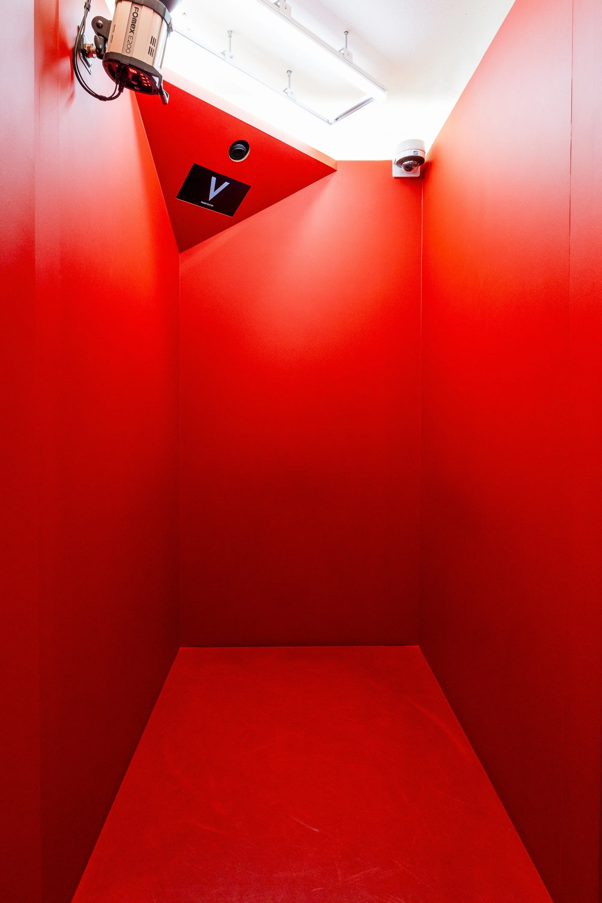 Small enclosed red room with a security camera, a V-shaped monitor, and a light fixture on the ceiling.