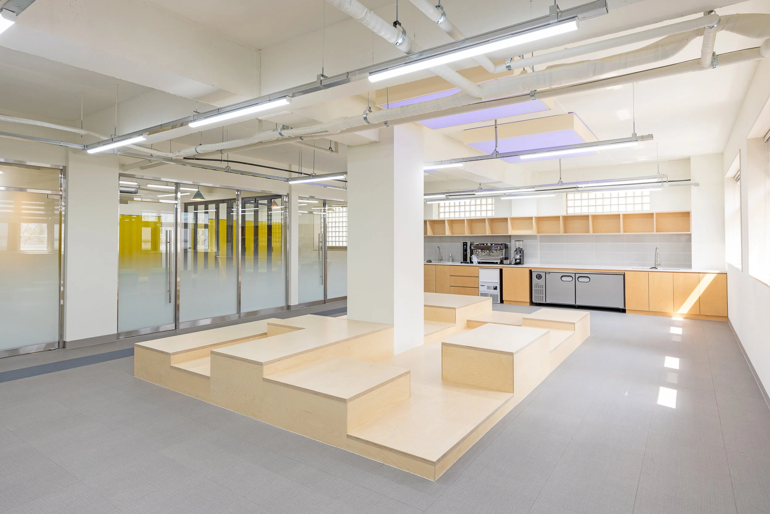 Modern open-concept office kitchen and lounge area with natural light, light wood cabinets, and gray flooring.