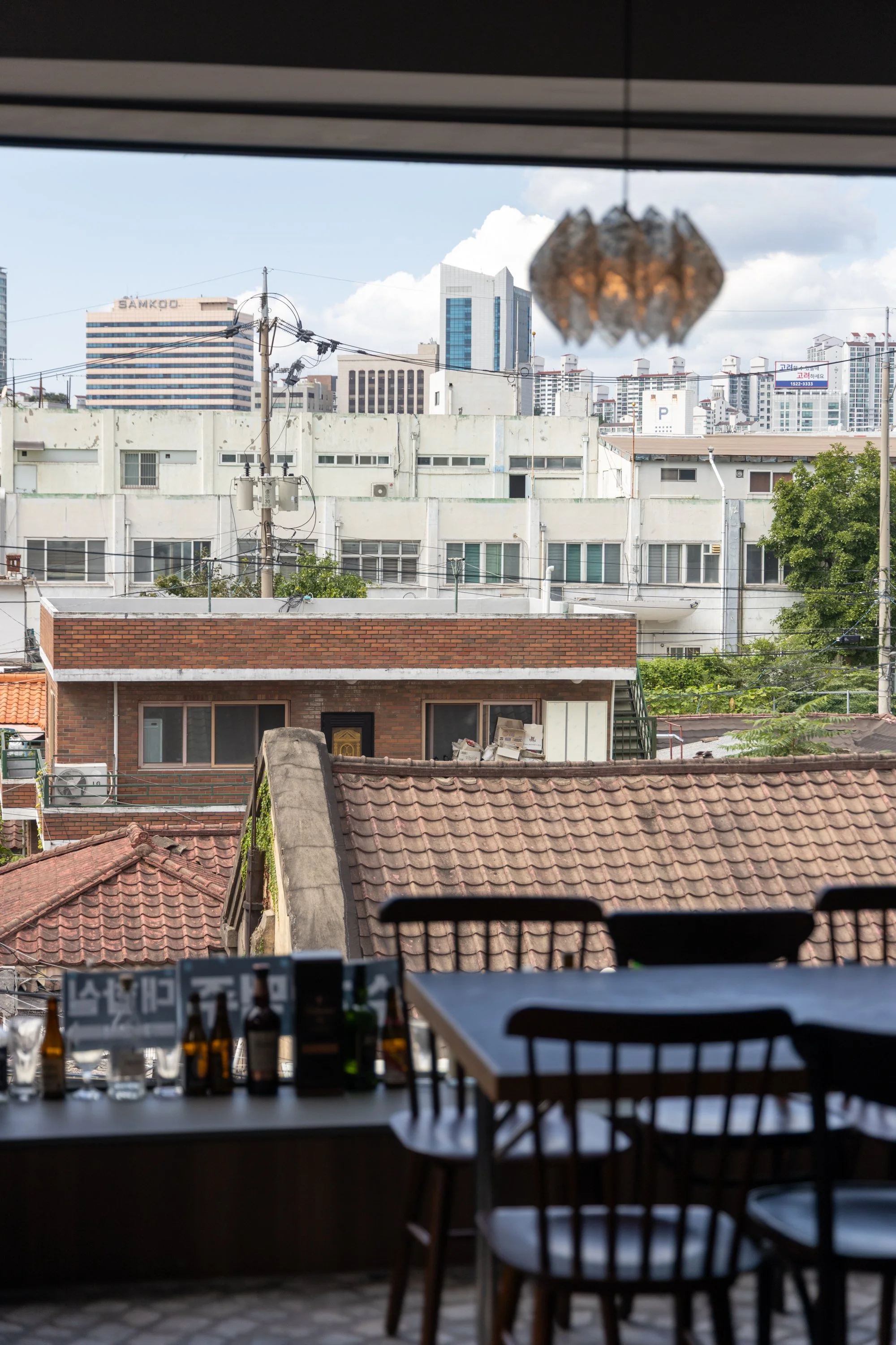 View of city buildings through a window, with a hanging lantern in the foreground and a table with bottles inside.