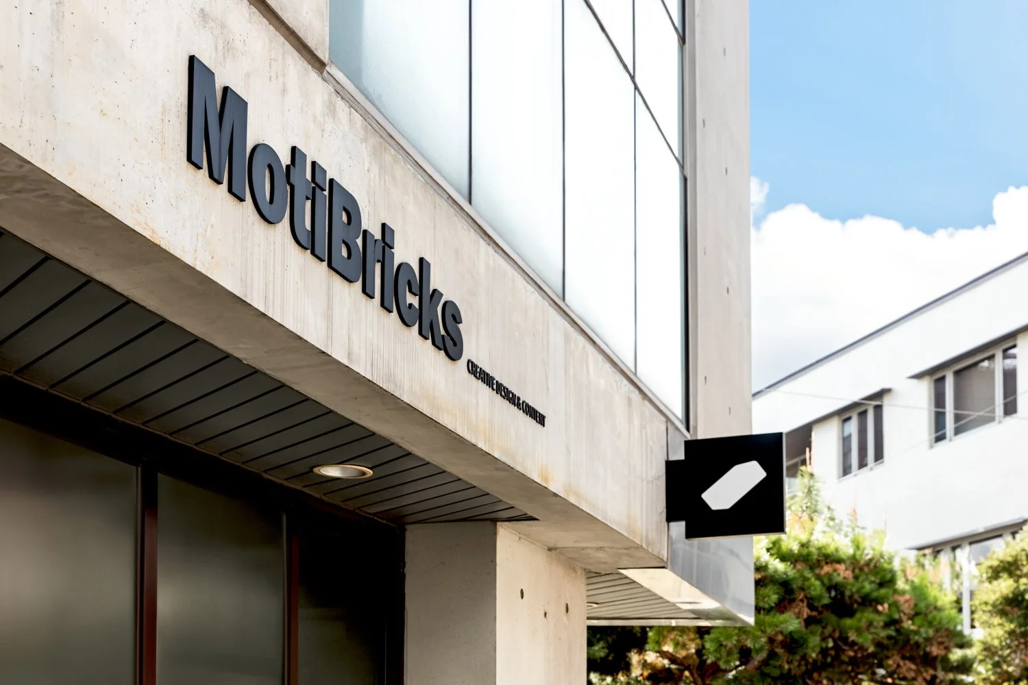 Exterior of a building with a sign that reads 'Motiblicks,' featuring large black lettering and a smaller tagline 'Creative Design & Content.' There is also a black square sign with a white shape, a tree with green foliage, and part of the building w