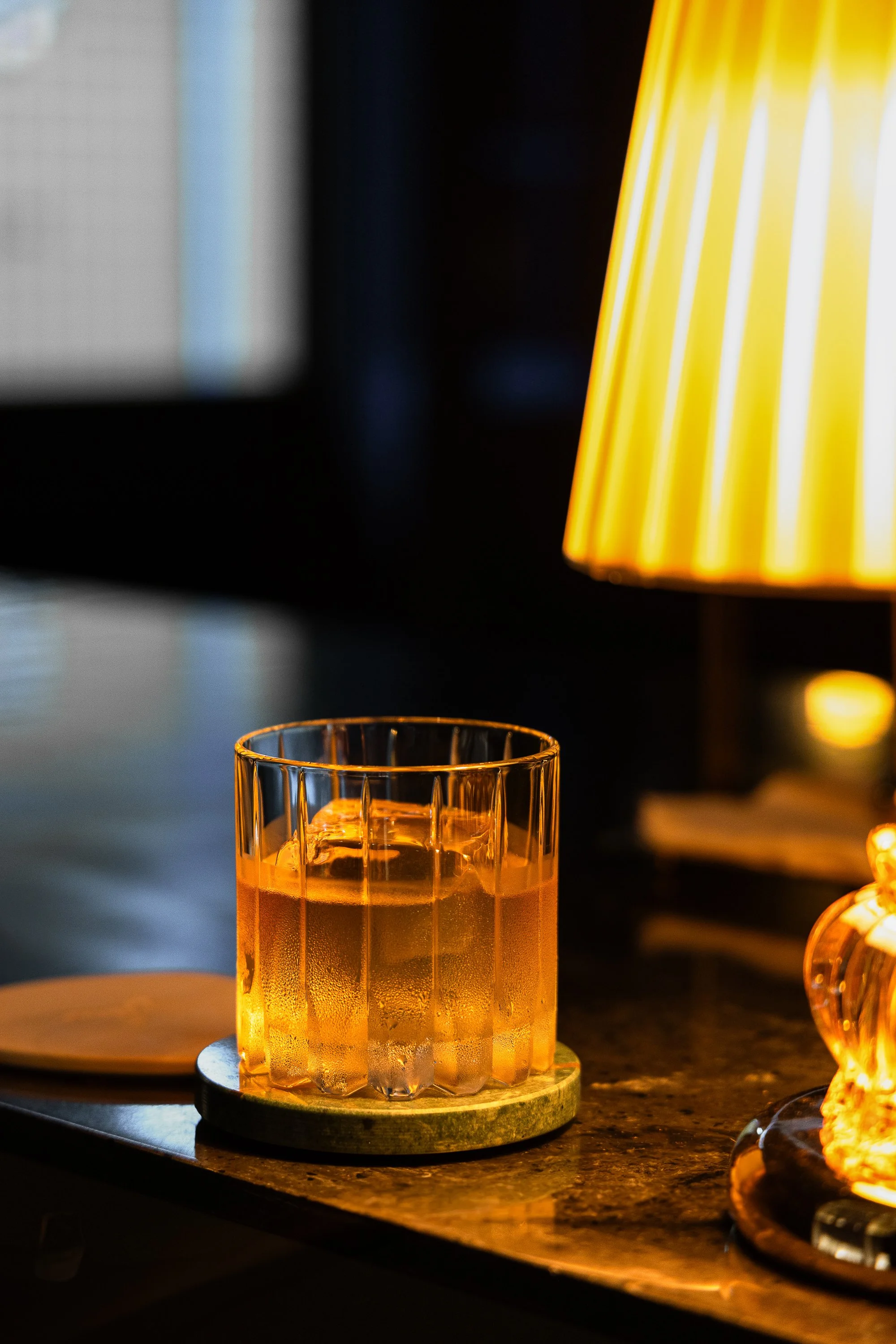 A glass of whiskey on a coaster placed on a dark countertop, with a yellow lampshade and blurred background.