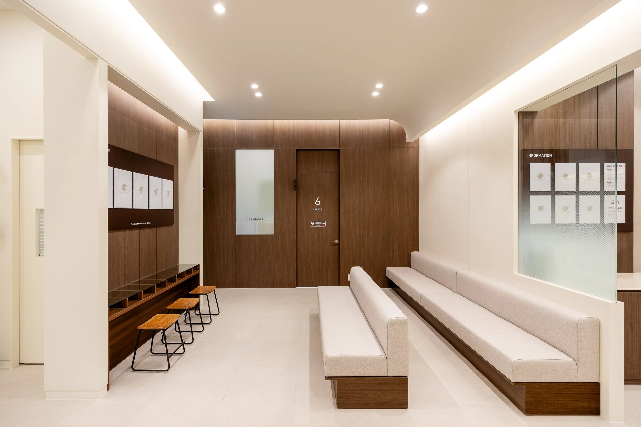 A modern waiting room with white walls, wooden accents, a long white bench along one wall, and four wooden stools in front of a wall-mounted display.