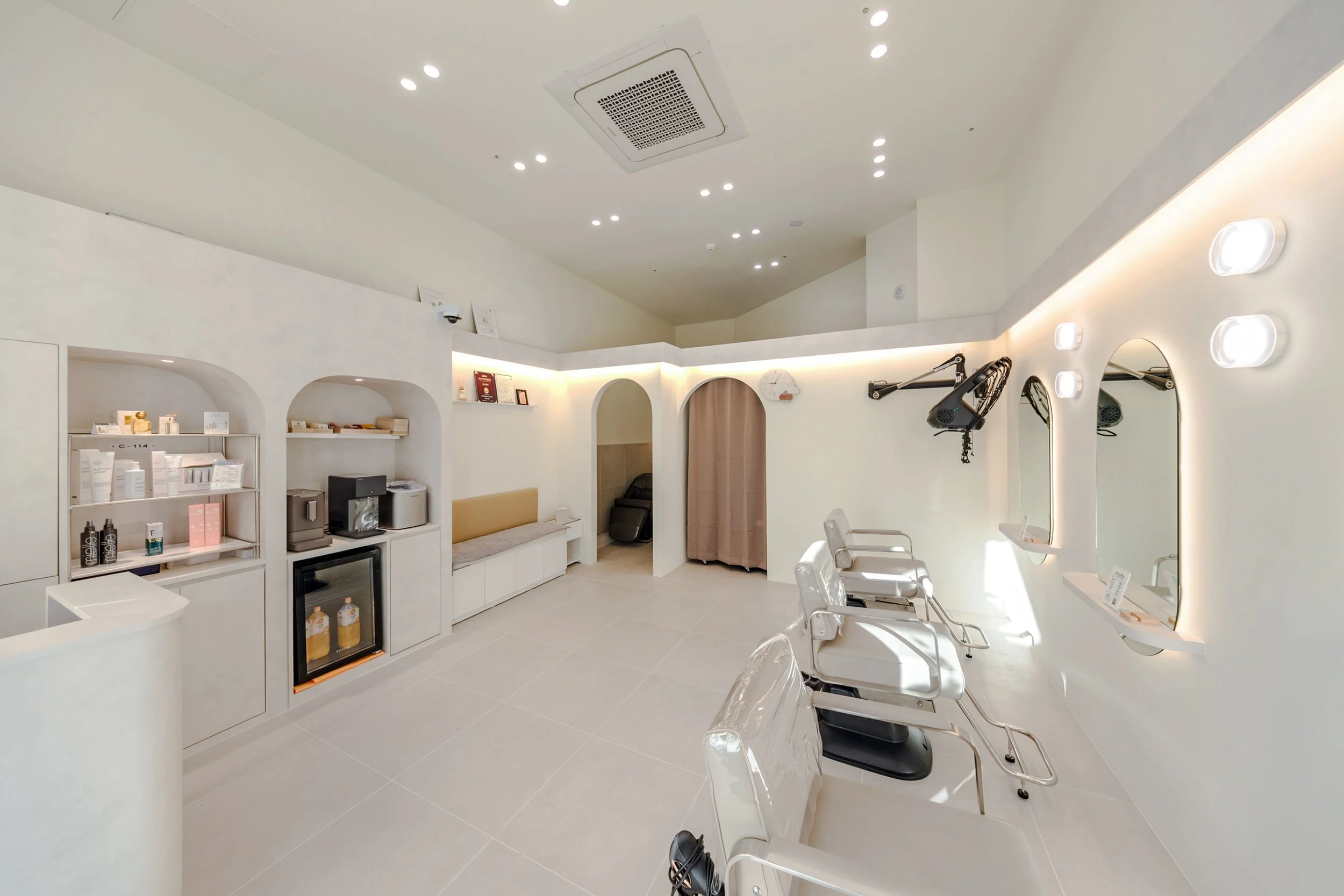 A modern, minimalist beauty salon or spa with white walls, three styling chairs in front of mirrors, and shelves stocked with hair and beauty products.