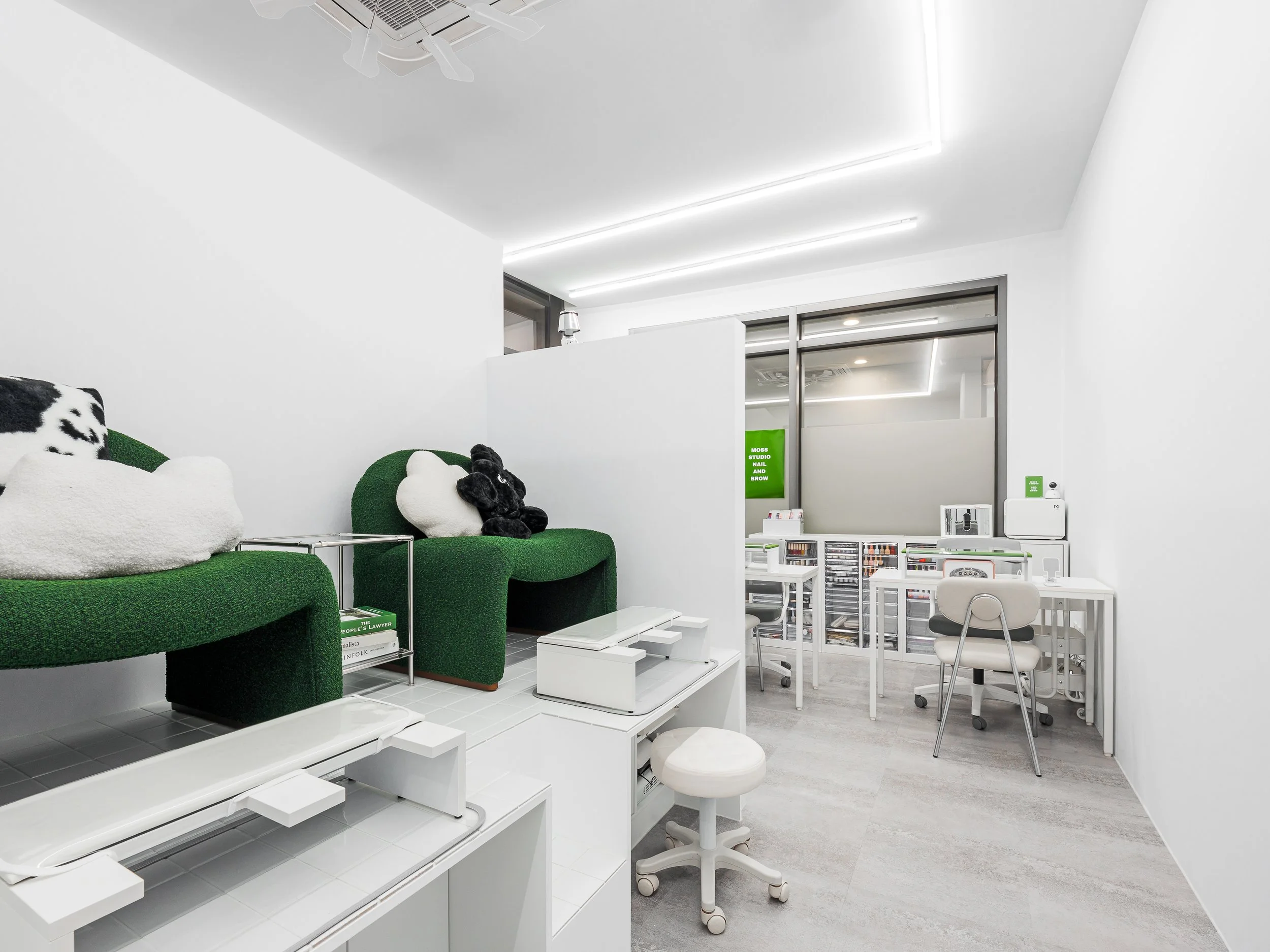 Modern, minimalist nail salon with white walls and furniture, green cushioned seating with plush animal pillows, and a sunlit workspace with nail polish displays and equipment.