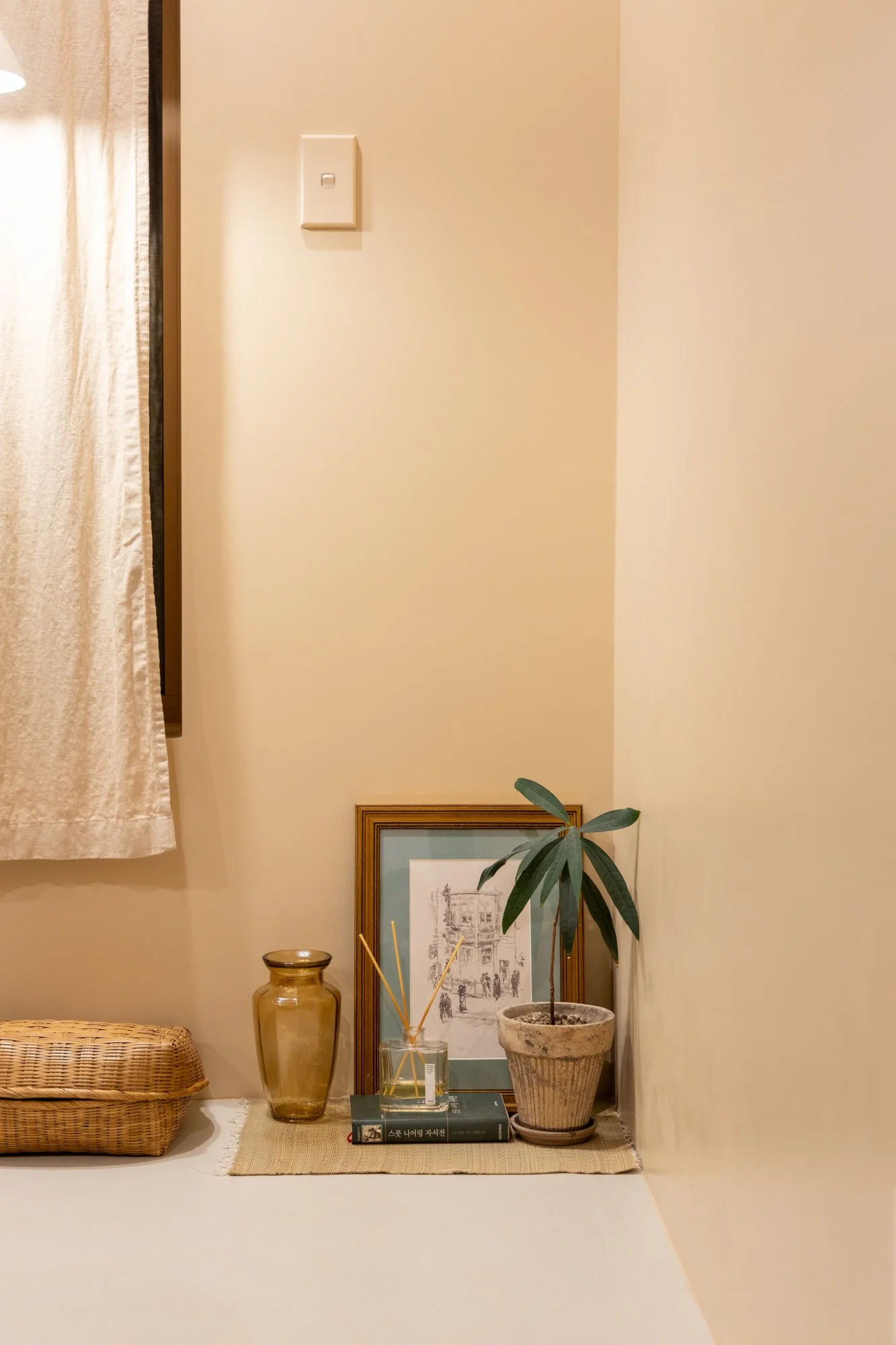 Decorative corner with a framed sketch, a potted plant, a book, a glass jar, and a woven basket on a beige table mat, against cream-colored walls.