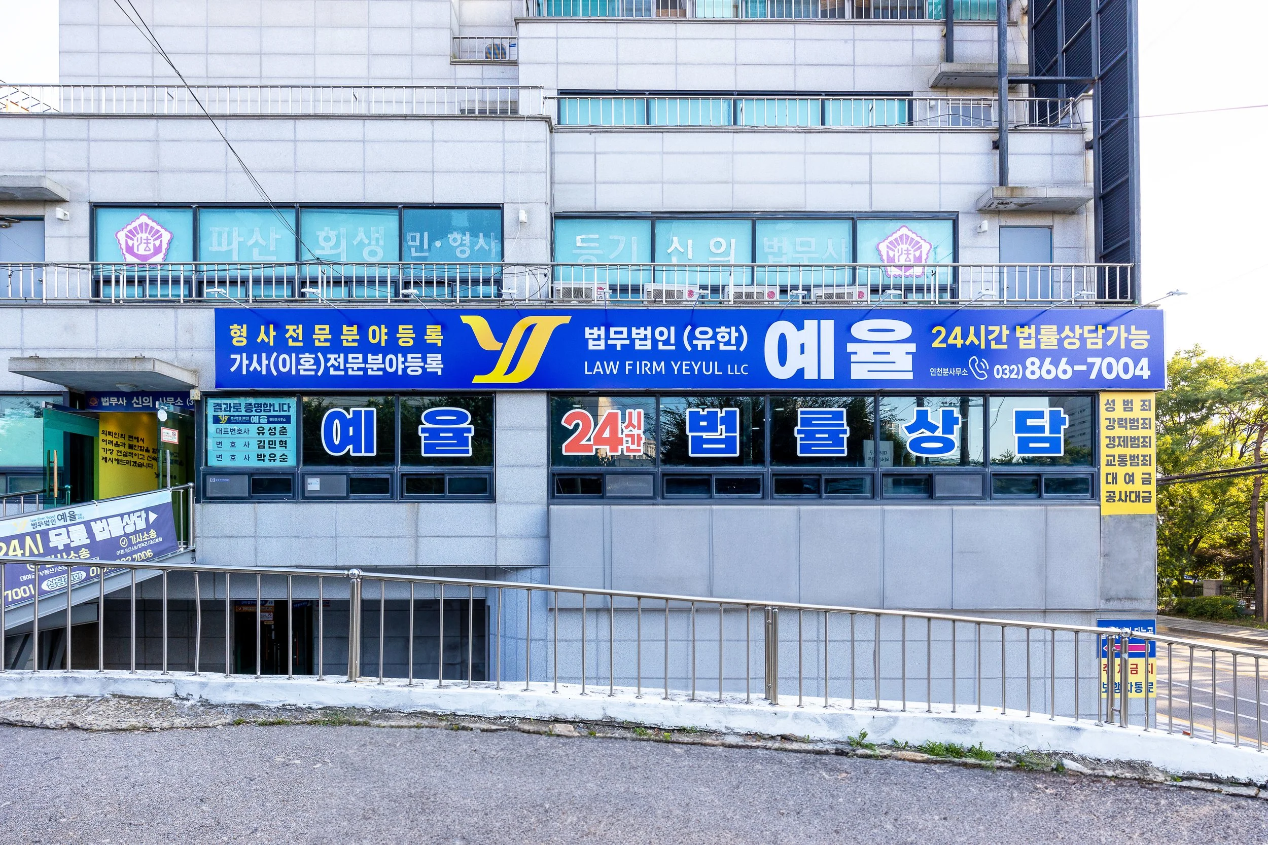 Exterior of a building with multiple blue and yellow signs advertising a law firm, including bilingual text in Korean and English, with a stairway leading down and a small yellow sign on the right side.