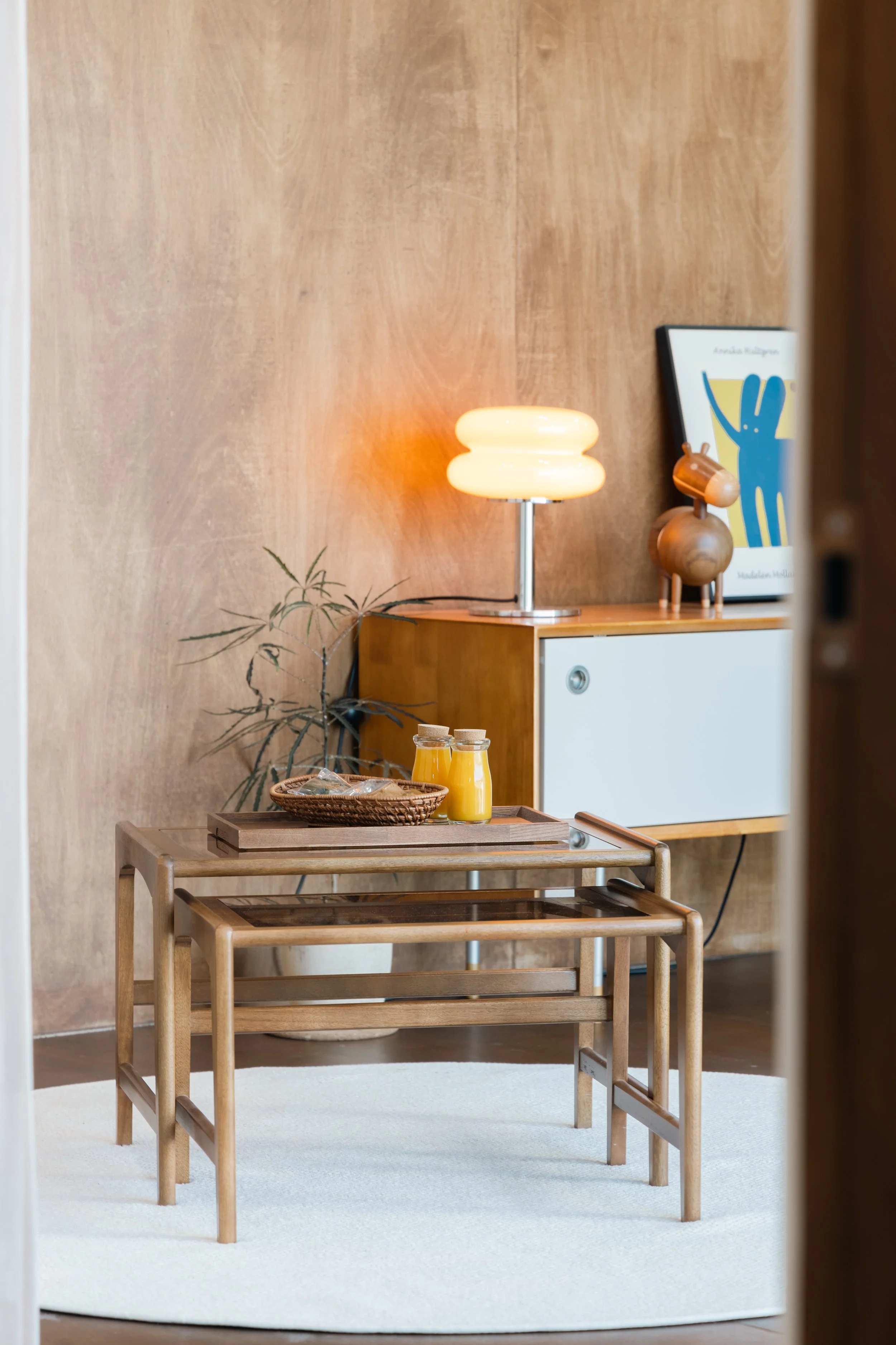 A modern living room corner with a wooden sideboard, a glowing lamp, a framed art print, a wooden dog sculpture, a potted plant, and a nested set of wooden tables with juice bottles and snacks on top, all on a white circular rug.