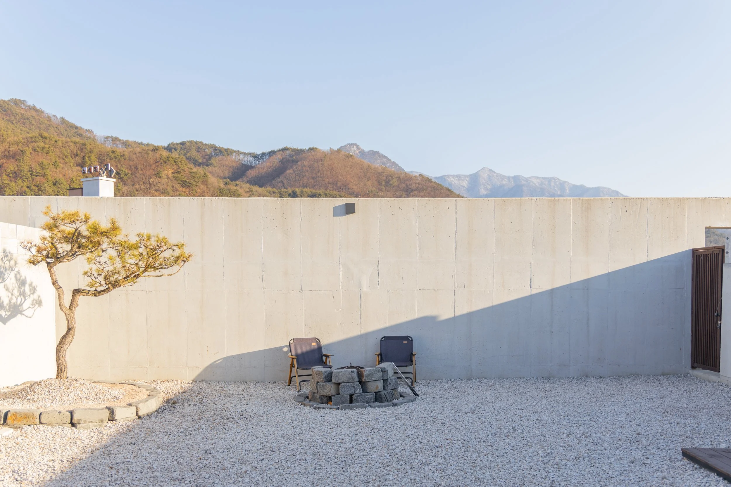 A minimalist outdoor patio with a small tree in a stone planter, two chairs with a circular stone fire pit, a high concrete wall, and mountains in the background under a clear sky.