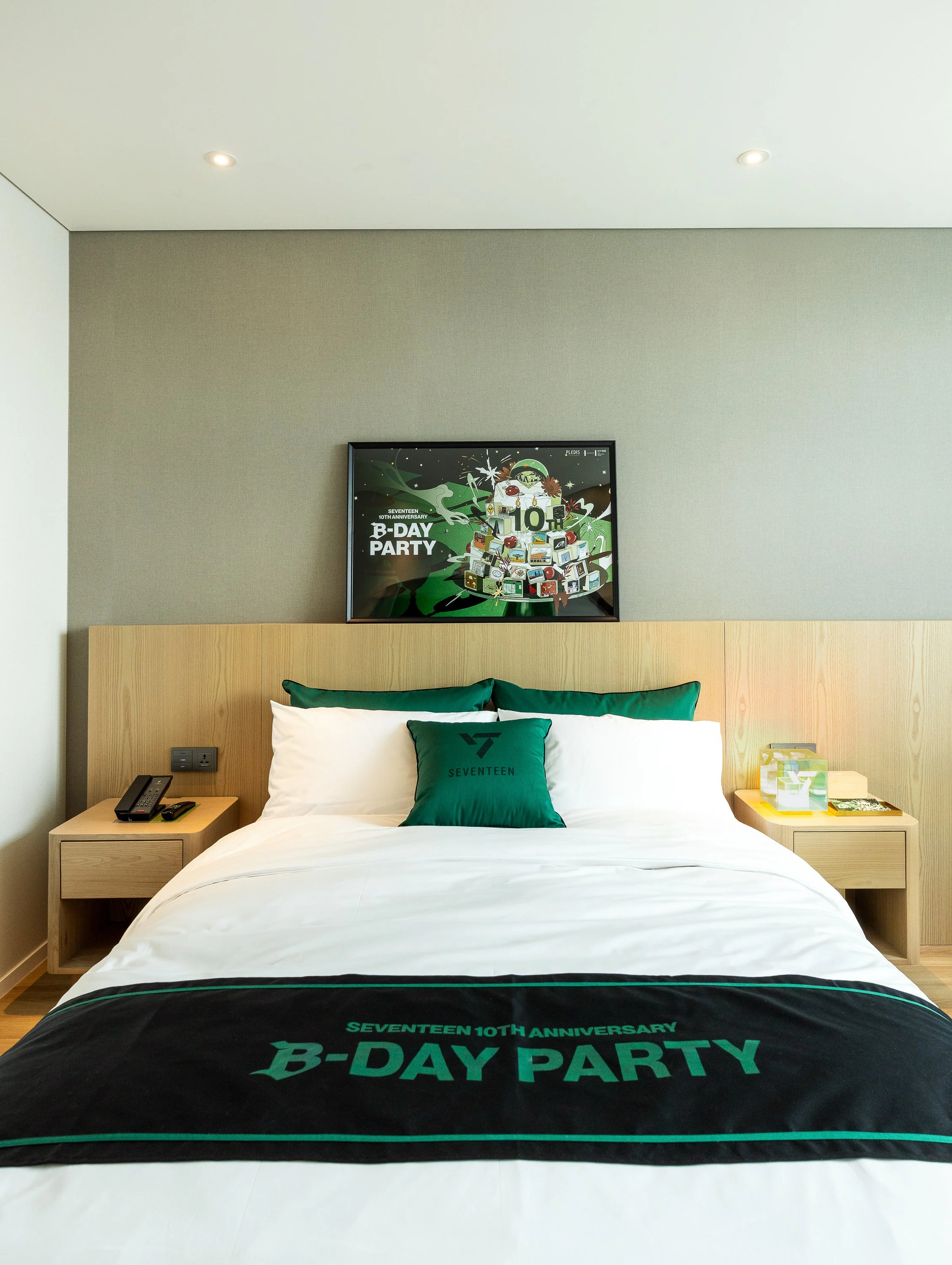 hotel room with a bed, green and white decor, a black blanket with green text, and a framed poster on the wall celebrating Seventeen's 10th anniversary and B-Day Party