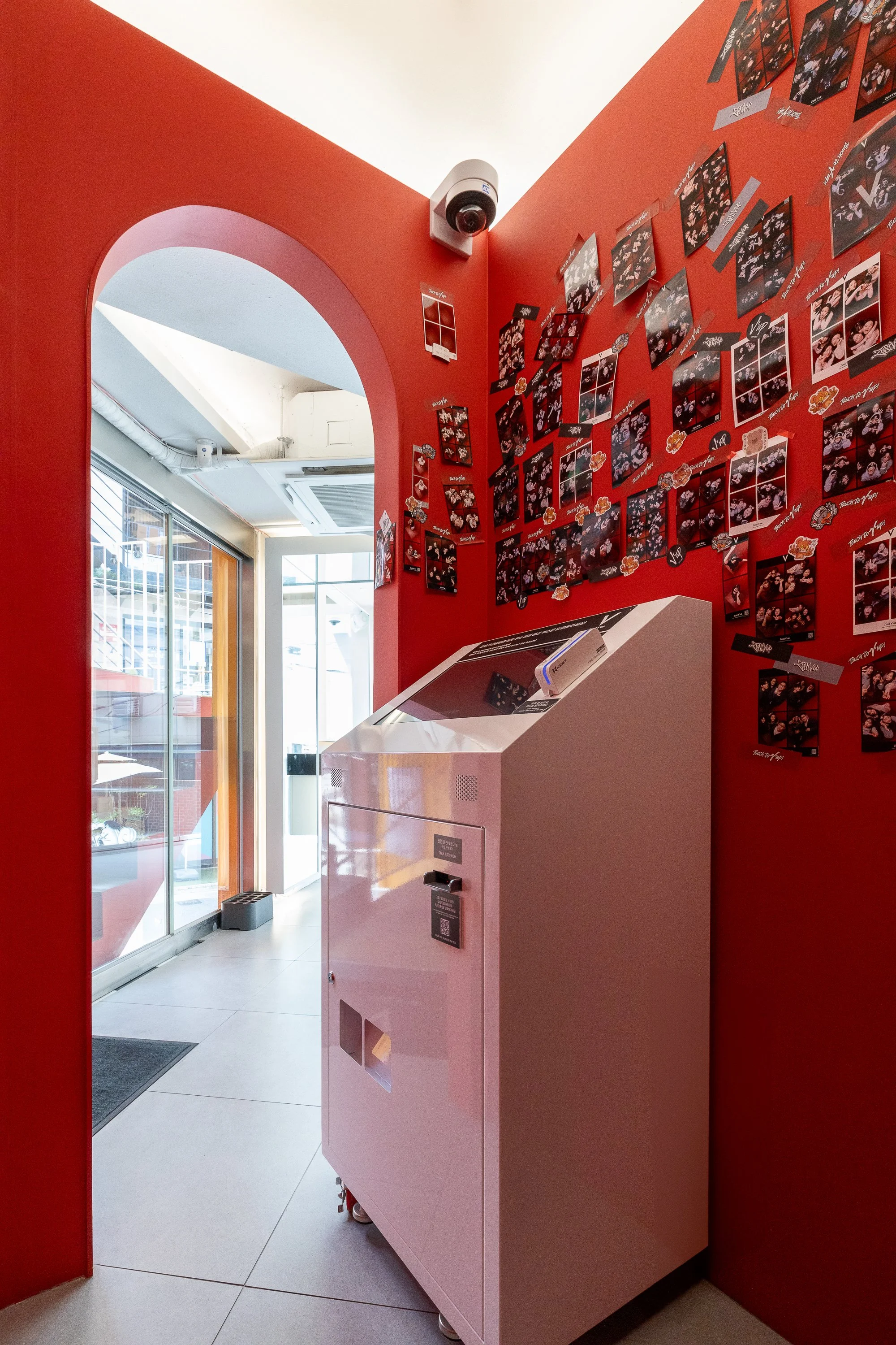 Photo booth machine against red wall adorned with numerous small photos and decorations, near glass door at entrance of building.