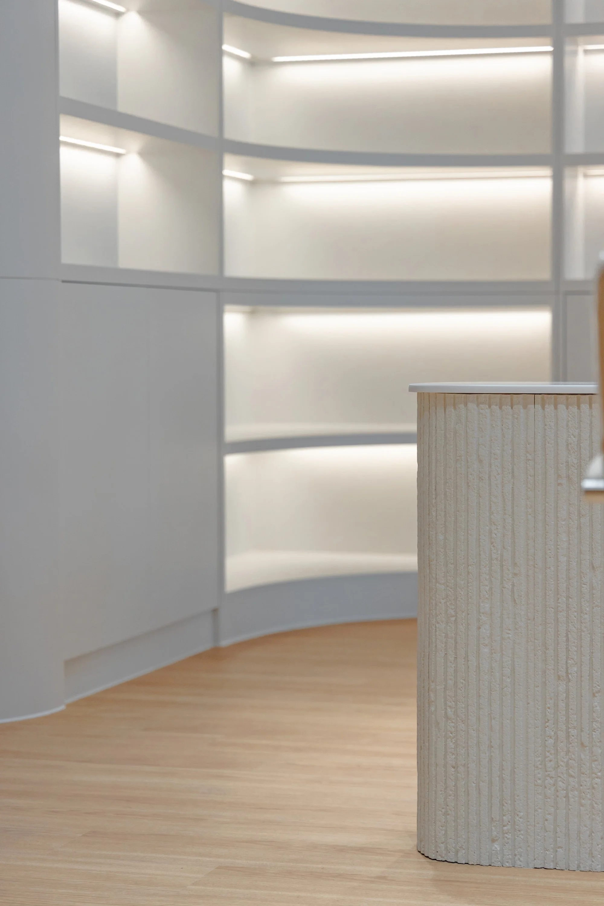 Modern, minimalist interior with curved white shelving and a textured beige cylindrical counter or column on a light wood floor.