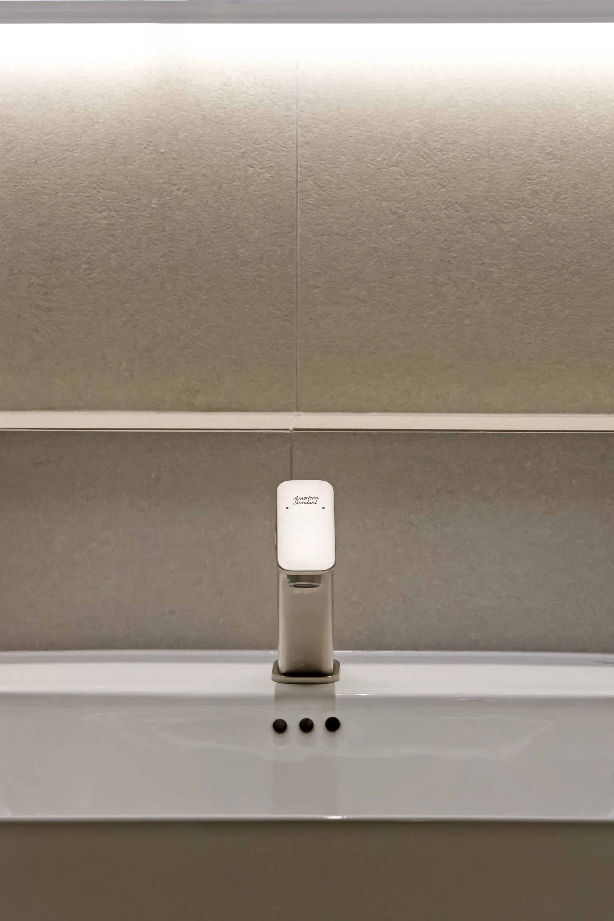 Close-up of a white American Standard hand sanitizer dispenser mounted on a beige tiled bathroom wall above a white sink with three black soap or sanitizer beads in front.
