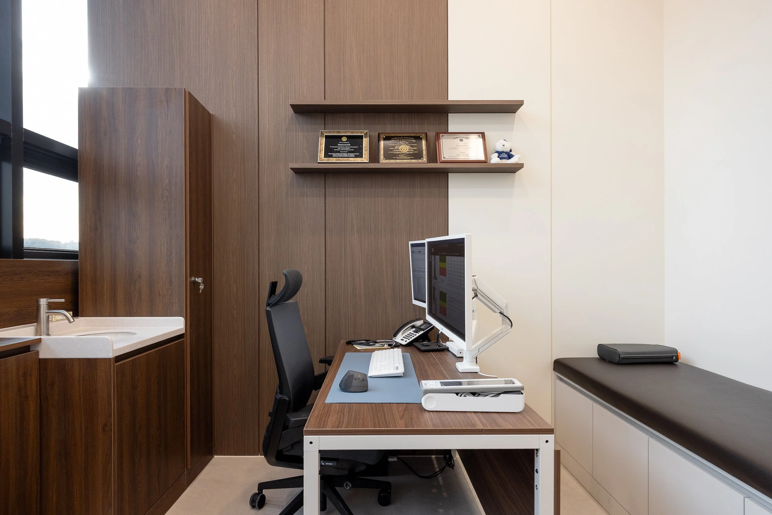 Medical office with a desk, computer monitors, and a patient examination bed along the wall, with framed certificates on a wood-paneled wall.