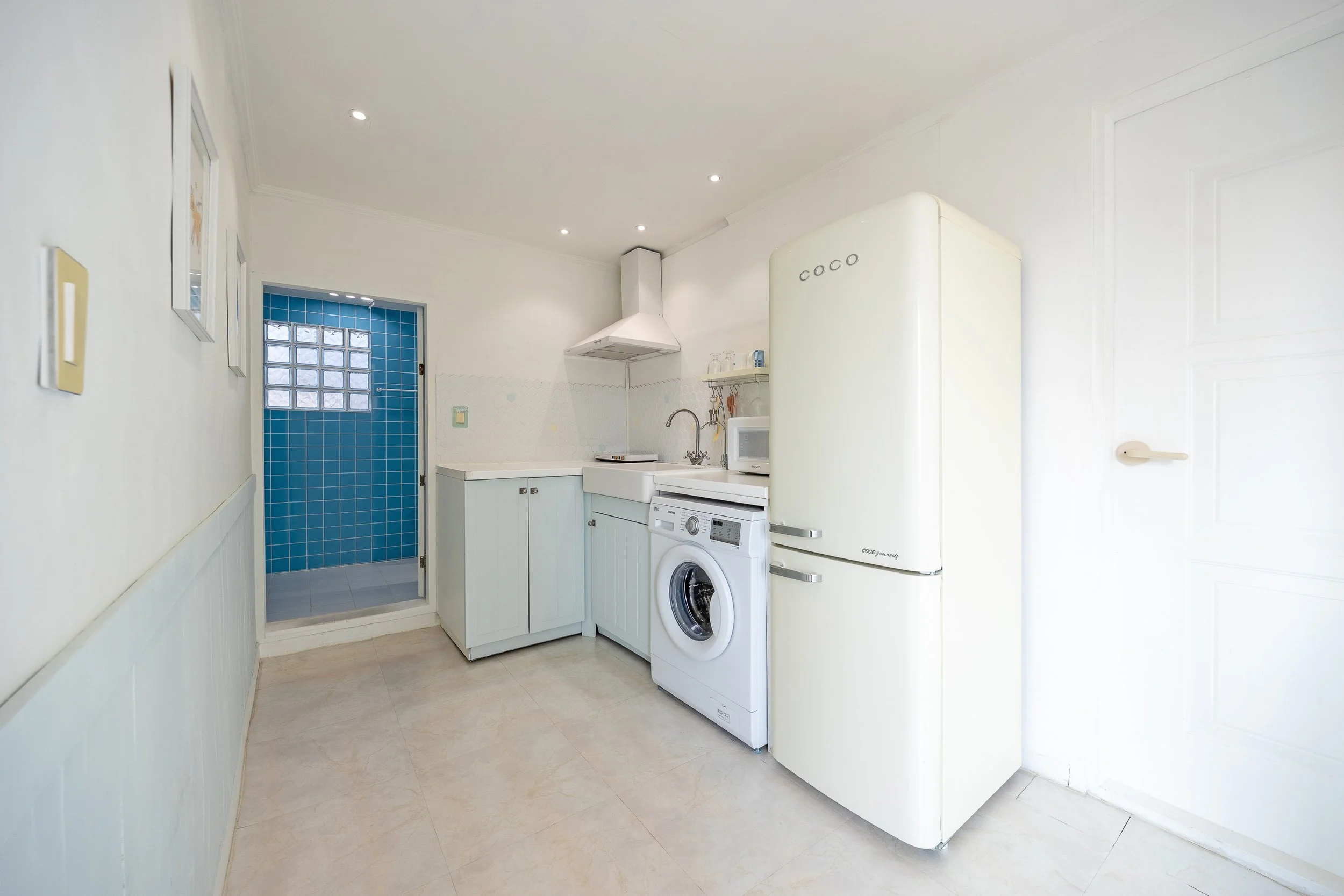 A small kitchen with a white refrigerator, washing machine, microwave, and light blue cabinets. There is a door leading to a shower with blue tiles and a glass block window.