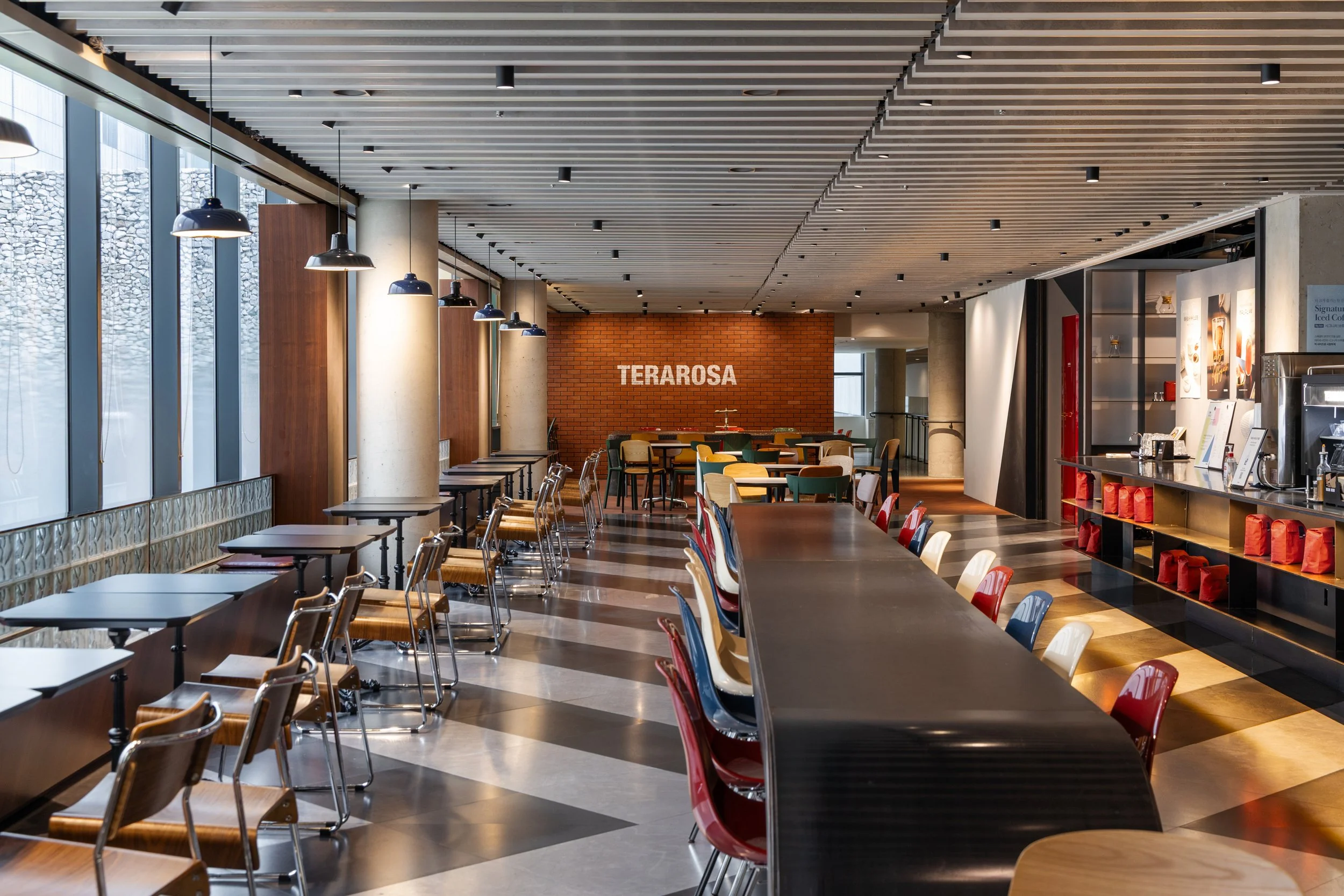 Modern, stylish interior of a café or restaurant with a long black table, colorful chairs, and a brick wall with the word "TERAROSA". Large windows on the left, pendant lights, and a food counter on the right.