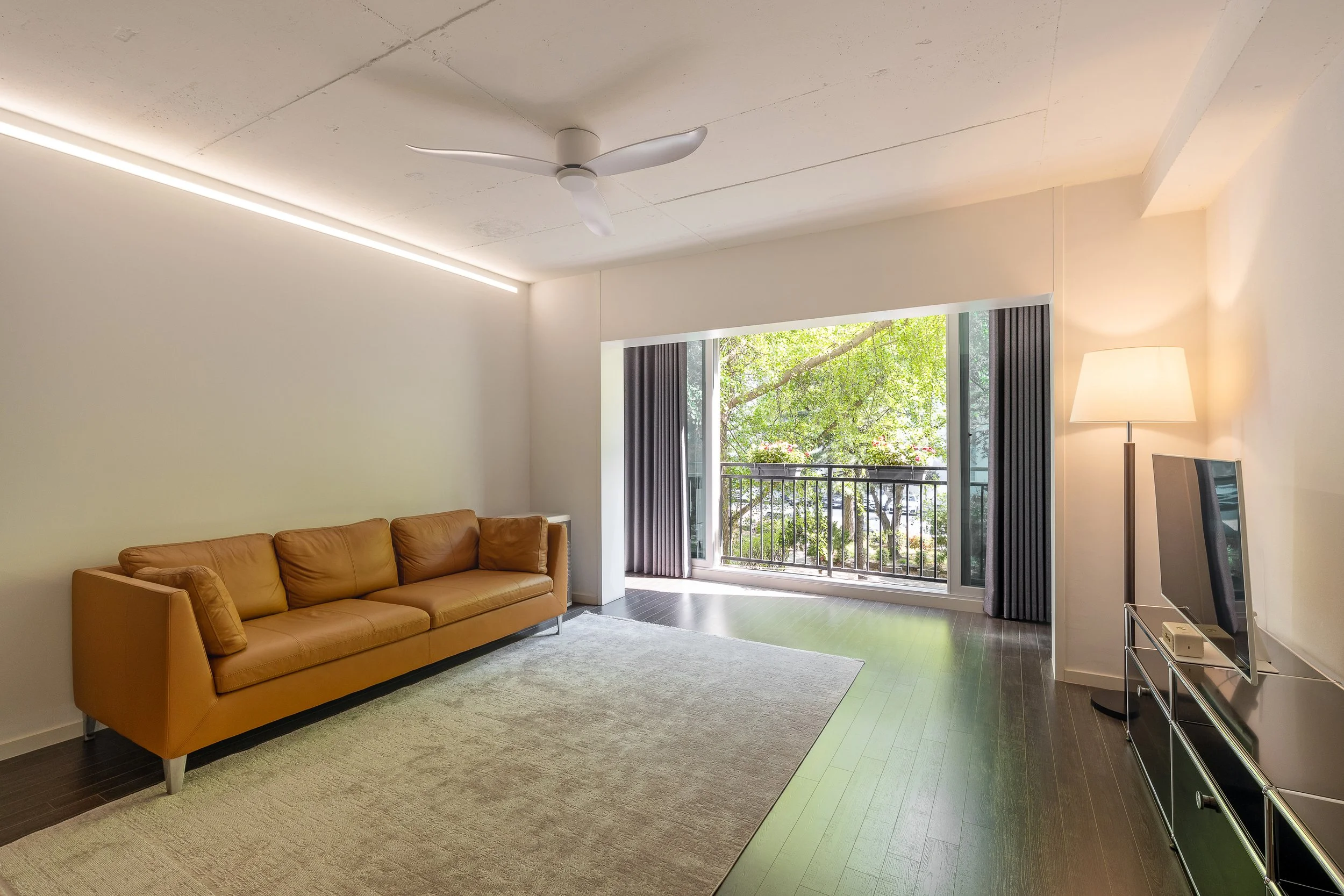 Living room with a tan leather sofa, a large window with a balcony and green trees outside, a white floor lamp, a flat-screen TV on a metal stand, a ceiling fan, and a beige area rug.