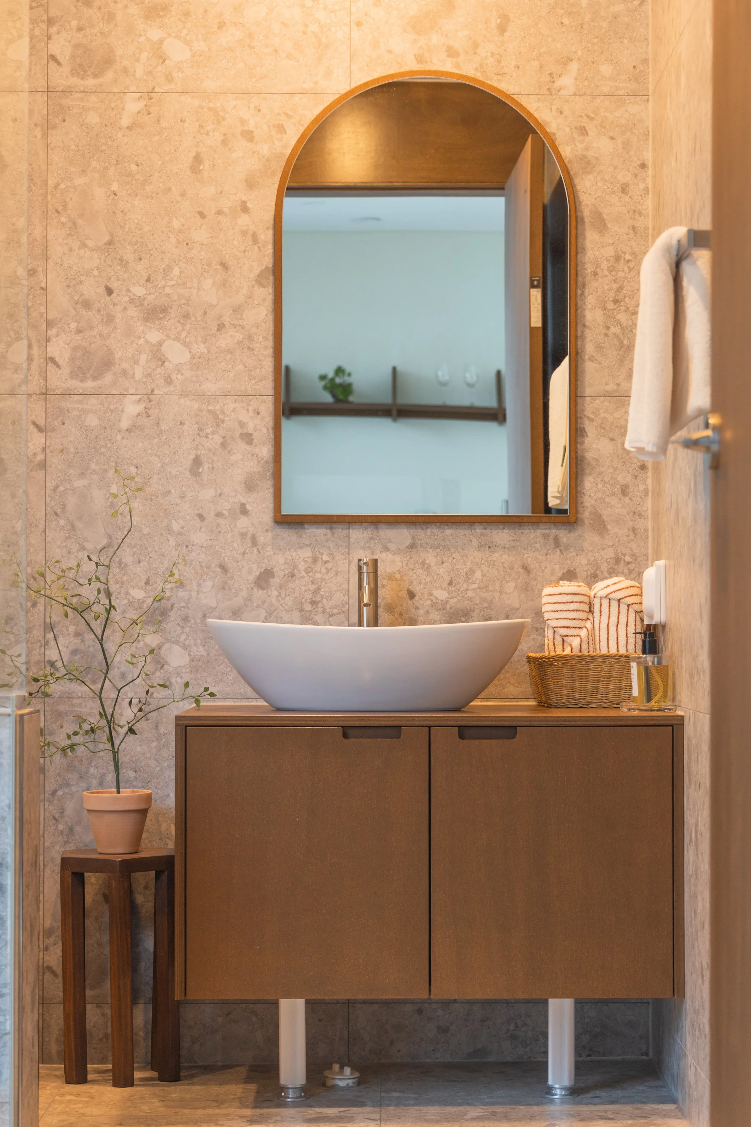 A modern bathroom with a wooden vanity, a white vessel sink, a round mirror above the sink, a small plant on a wooden stool, and a basket with rolled towels on the counter.