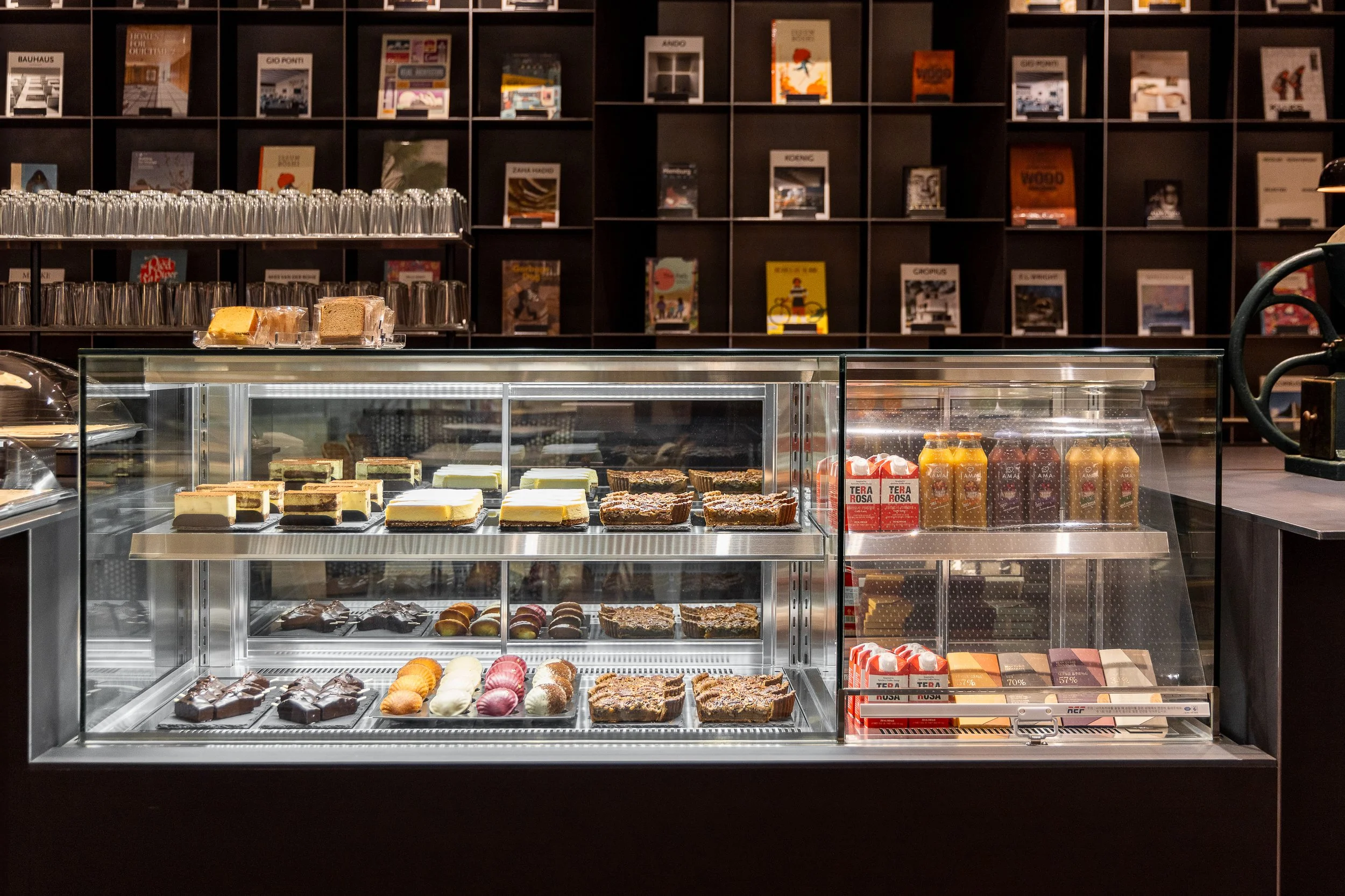 Display case with assorted cakes, pastries, and chocolates, with bottled drinks on the side, in a café or bakery setting. Shelves with books or magazines are in the background.