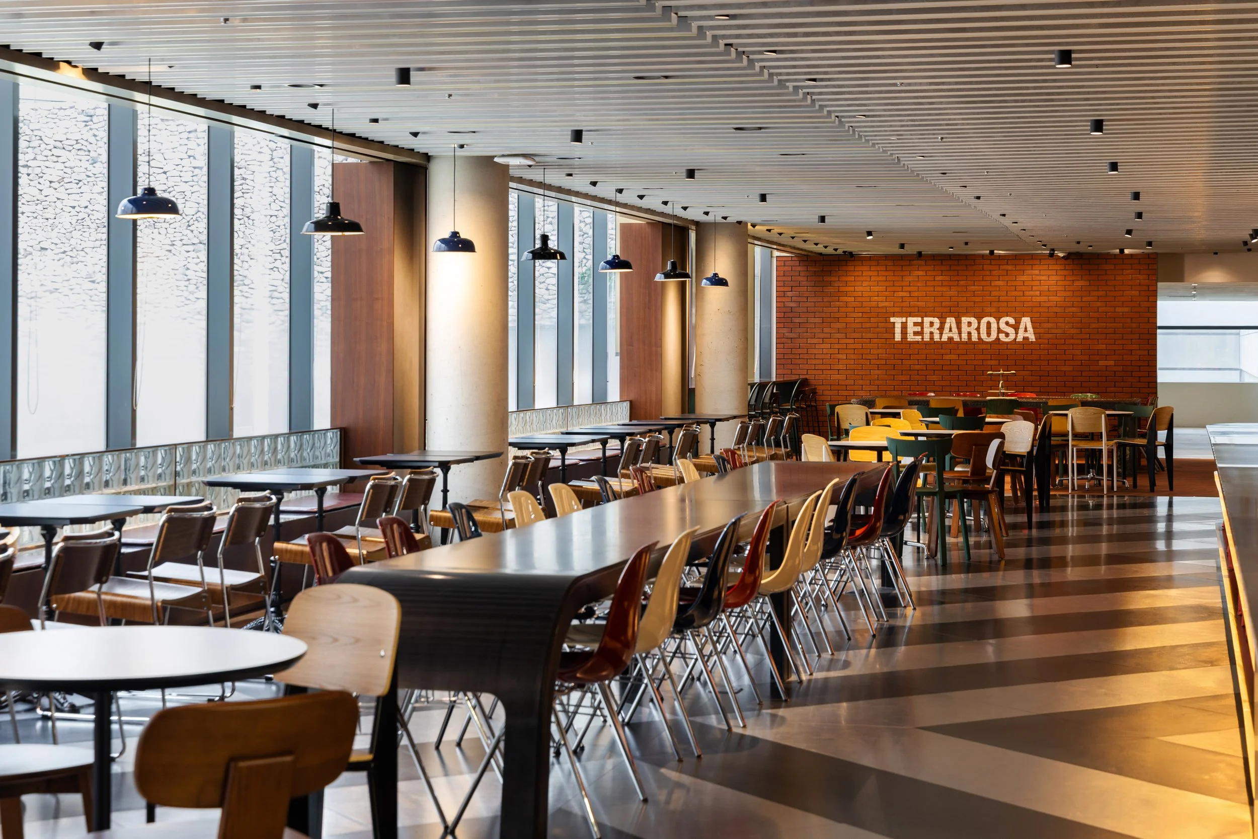 Interior of a modern restaurant or cafe with a long shared table, various smaller tables and chairs, large windows, pendant lighting, a brick wall with the word 'TERAROSA' displayed, and a wooden and brick decor.