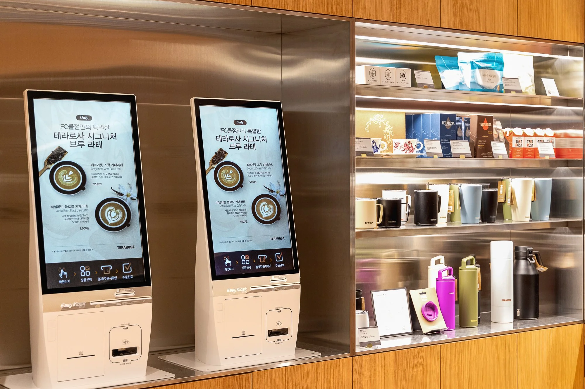 Two digital kiosk screens displaying coffee menu options in Korean, set against a display of coffee mugs, tumblers, and packaged coffee products on shelves in a coffee shop or cafe.