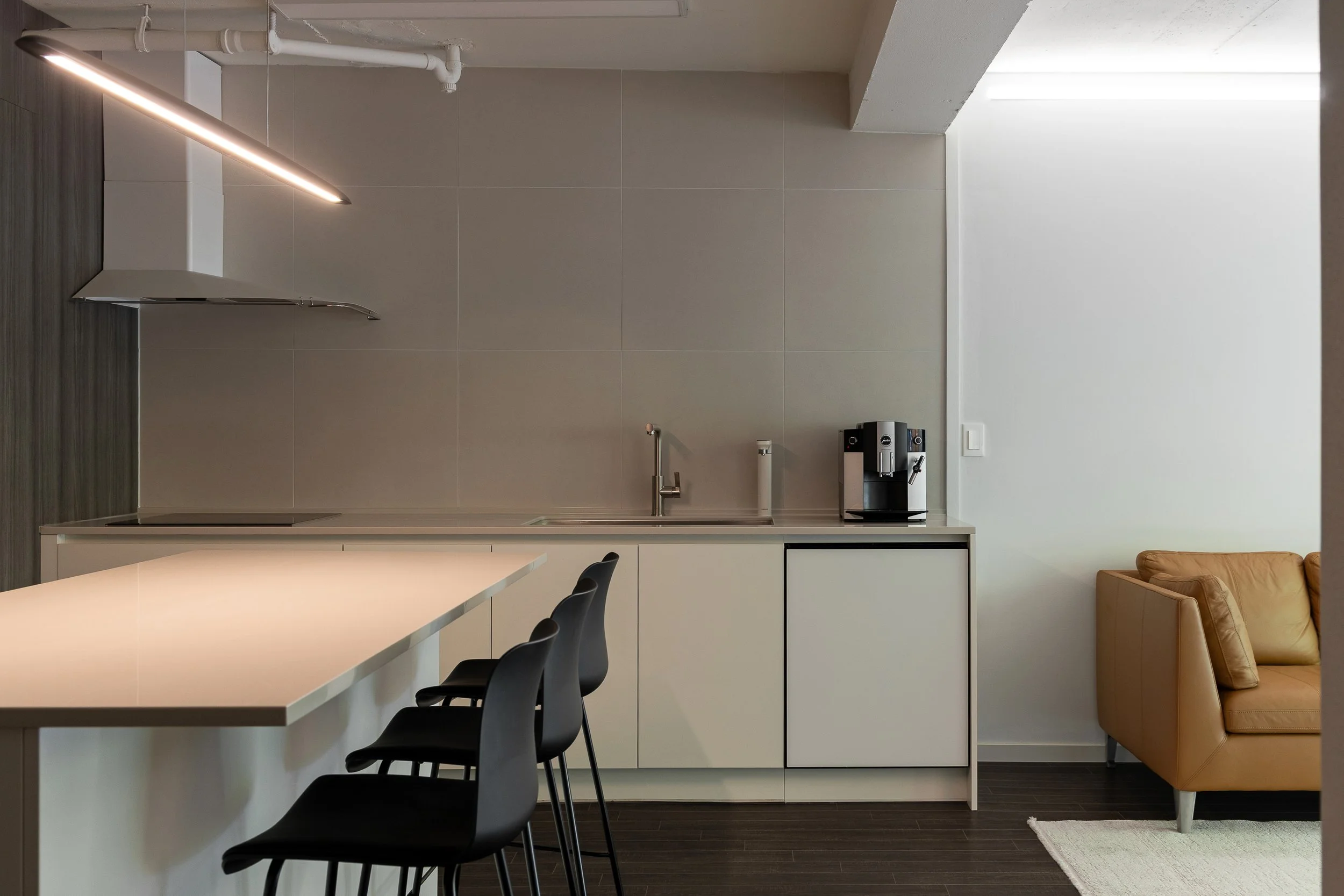 Modern kitchen with white cabinetry, black barstools, beige sofa, and coffee machine.