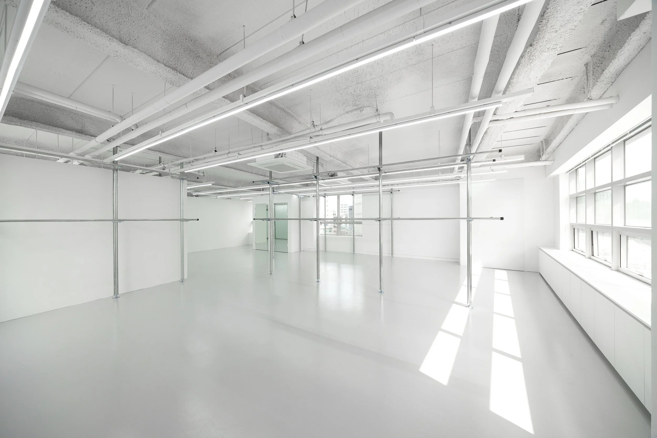 Empty, white interior space with large windows, overhead lights, and scaffolding structures, resembling a modern, minimalist studio or office.