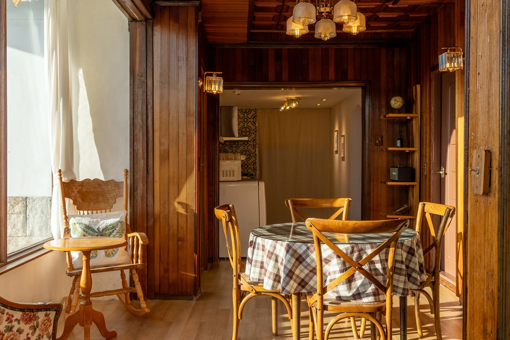A cozy dining area with a round, checkered tablecloth on a wooden table, surrounded by four wooden chairs, in a room with wood-paneled walls and a large window with white curtains, sunshine streaming in.