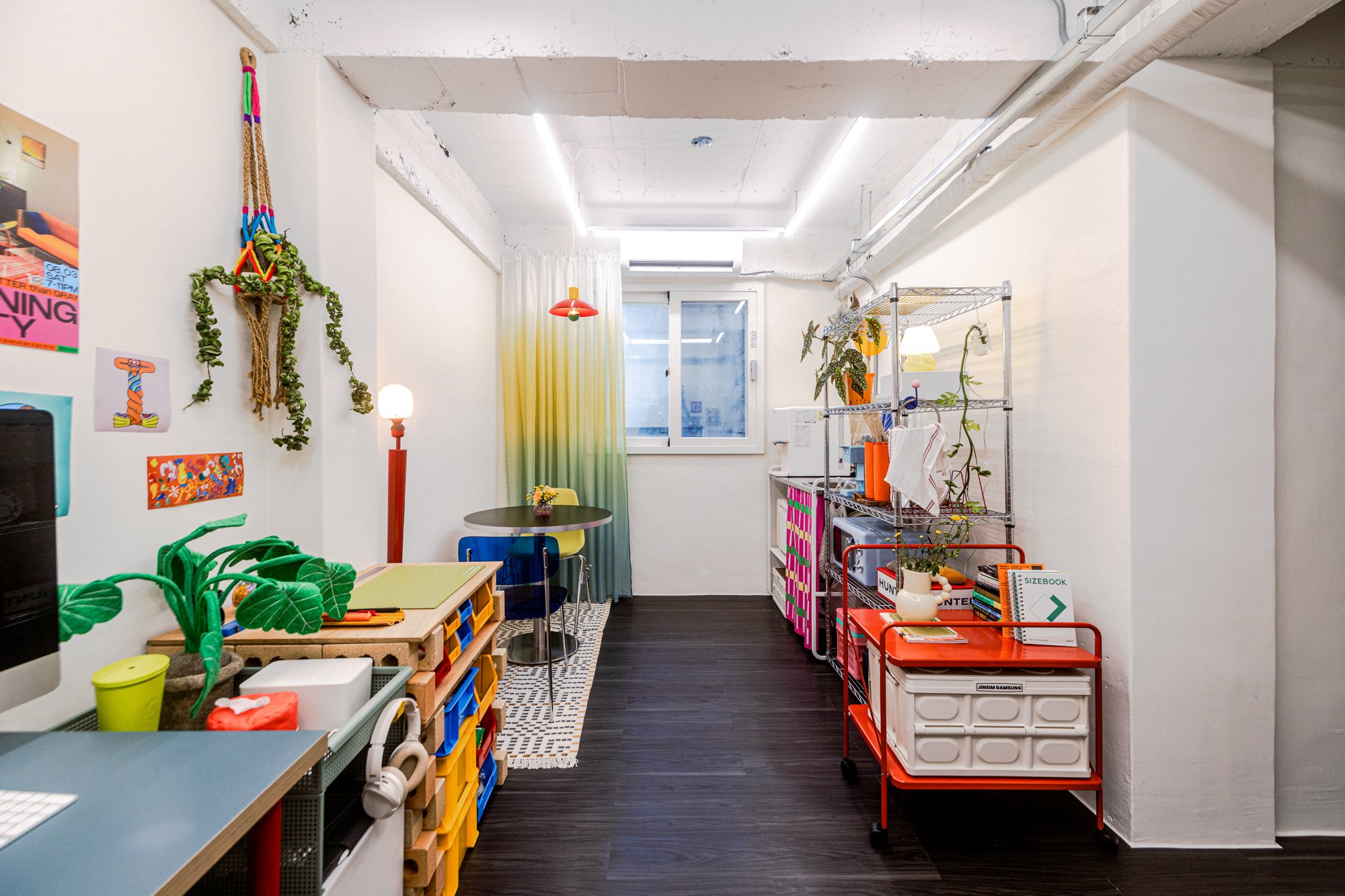 Colorful interior space with furniture, plants, posters, and decorative items, including a window with yellow-green curtains.
