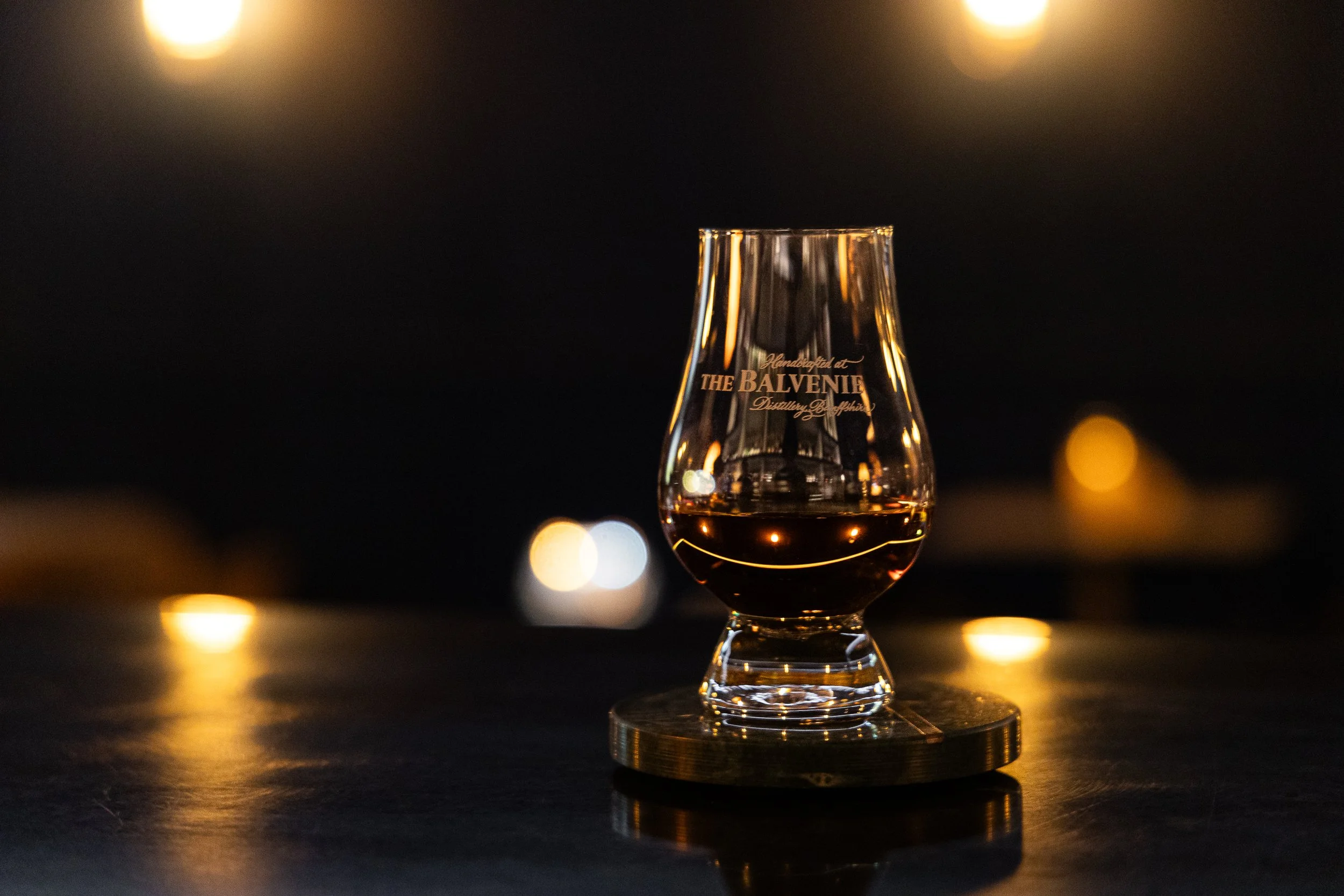 A glass of dark liquor on a round coaster, illuminated by warm lighting, with a blurred dark background and bokeh lights.