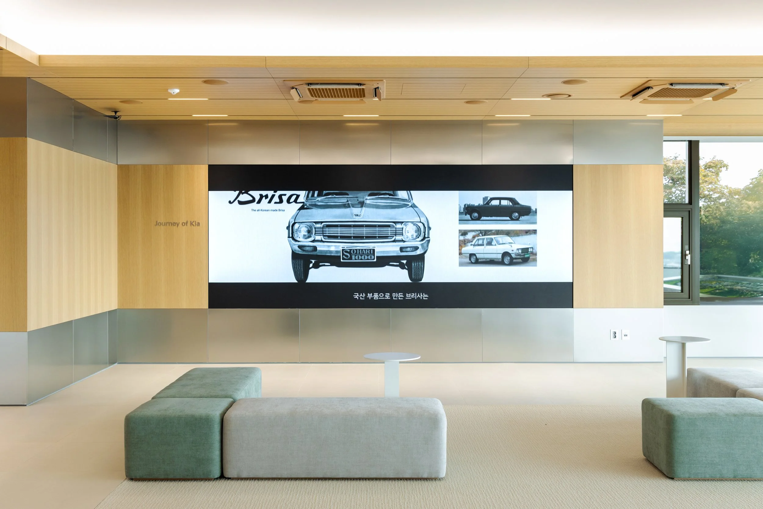 Modern lounge with a large digital screen displaying images of vintage Kia cars, light wood and metal interior, two small white tables, and seating area with green and beige cushions, next to large windows showing outside greenery.