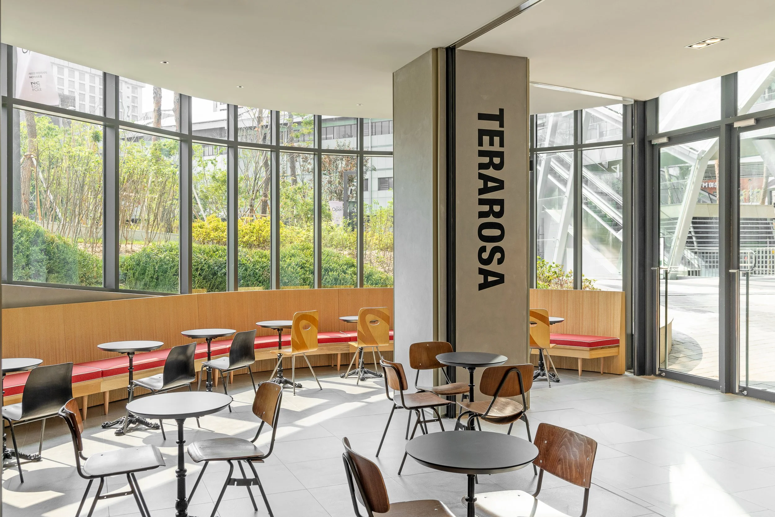 Interior of a modern café with round black tables, wooden and black chairs, and a curved wooden bench along large glass windows overlooking green outdoor plants and trees. There is a concrete pillar with the word "TERRA ROSA" written vertically.