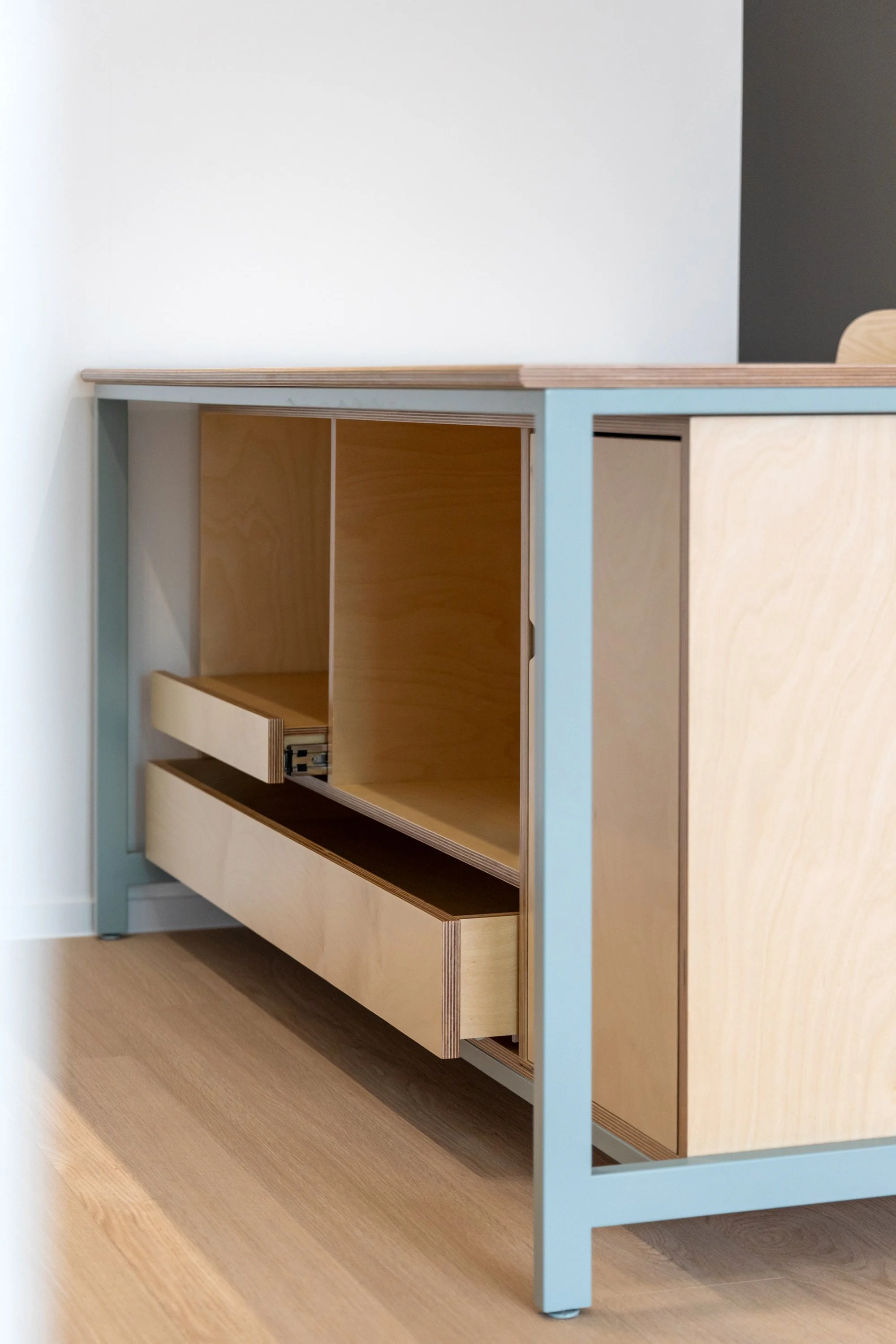 A modern wooden cabinet with blue metal frame, partially open drawer, and sliding doors, set on a hardwood floor.
